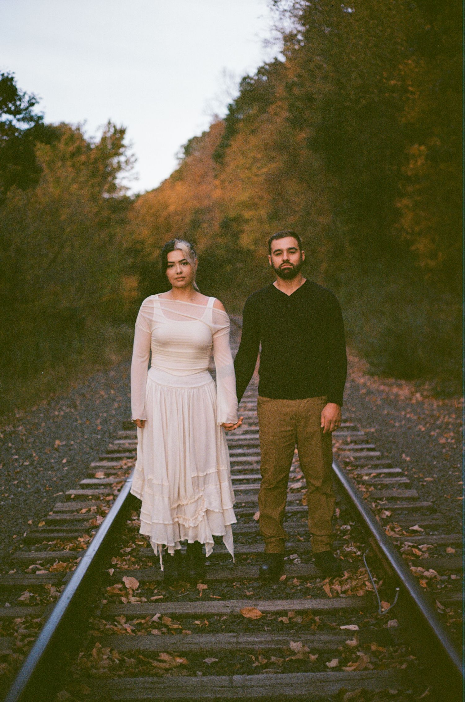 A couple stands together on train tracks surrounded by fallen autumn leaves in a romantic outdoor setting.