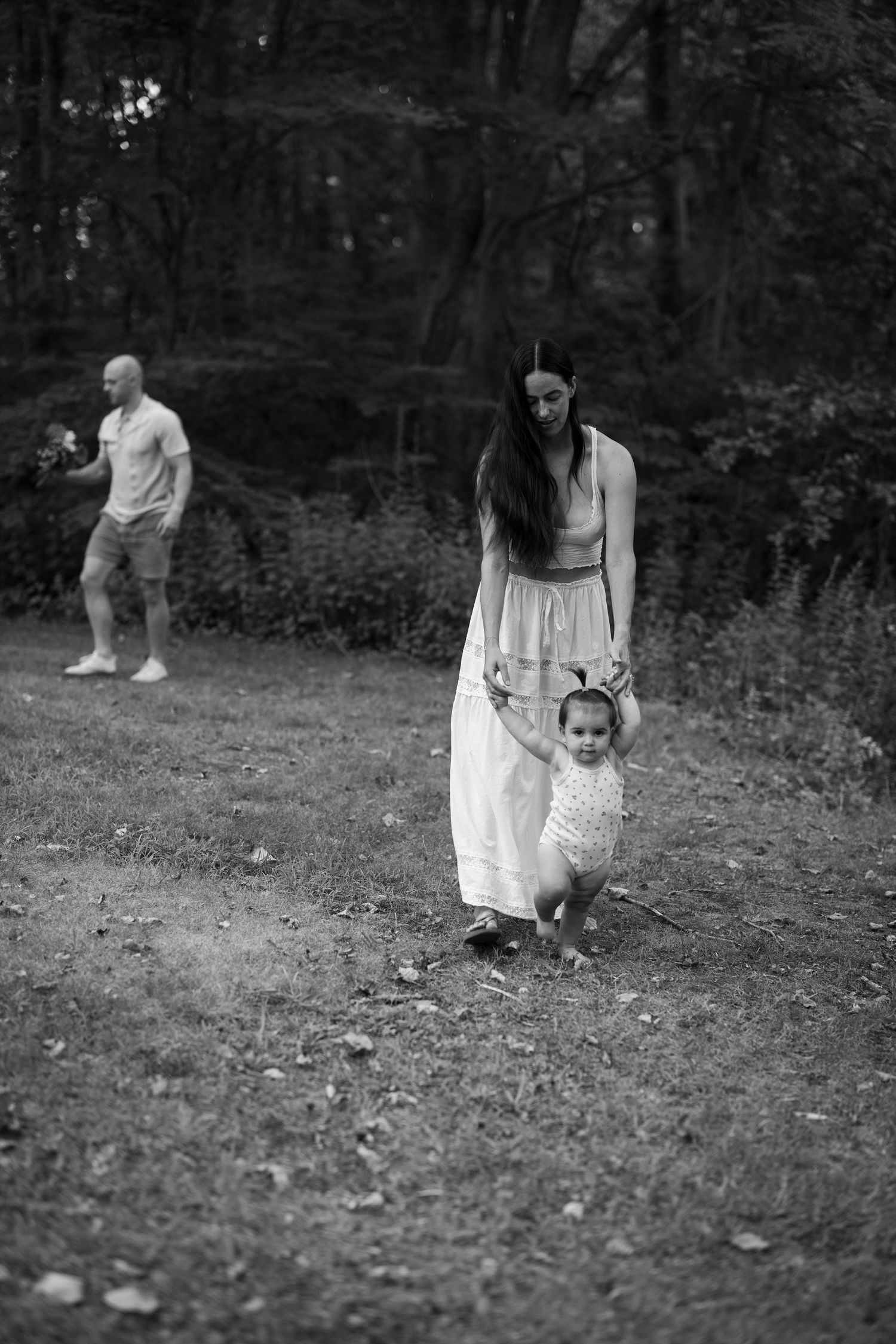 black and white candid family portrait. Family walks around the park. Mother holds toddlers hands as she walks.