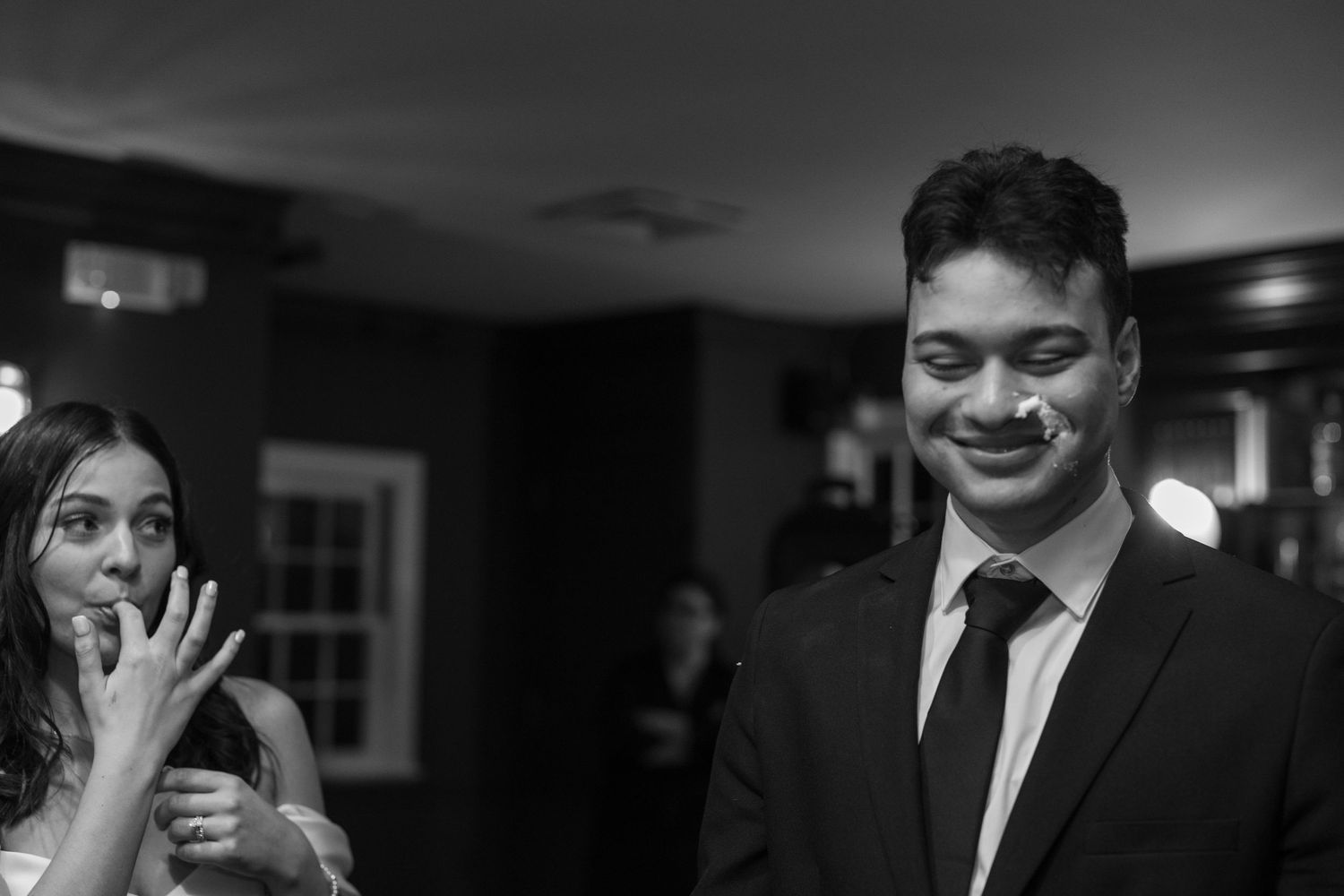 Black and white photo of a smiling man in a suit with cake on his face at a wedding reception.