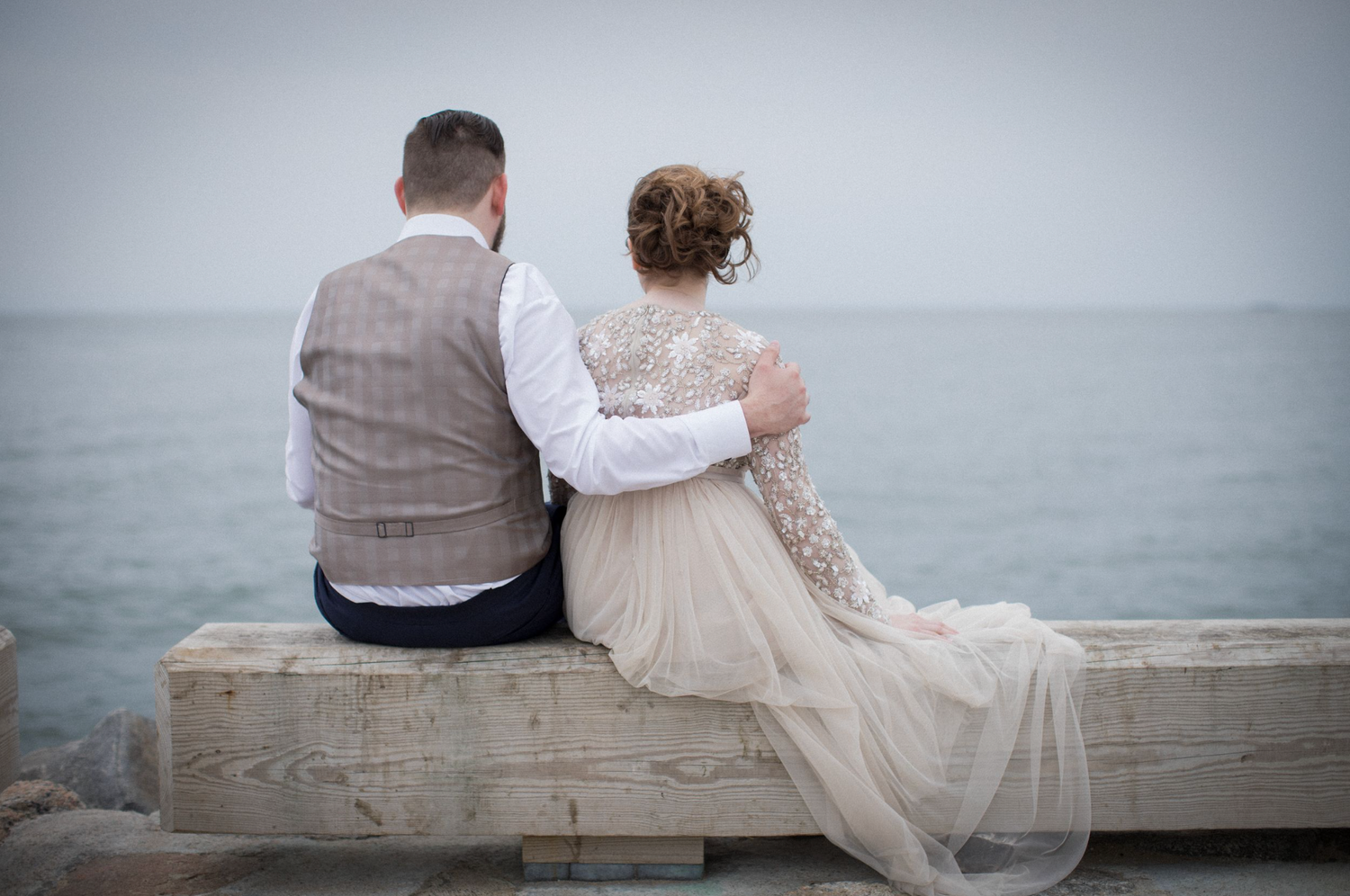 A couple sits together on a stone ledge overlooking a misty ocean in a romantic seaside moment.