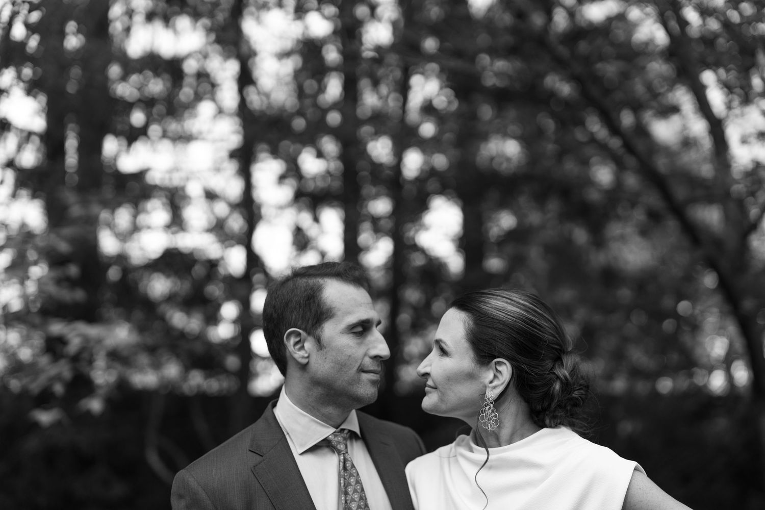 A black and white portrait of a couple gazing at each other outdoors with trees in the background.