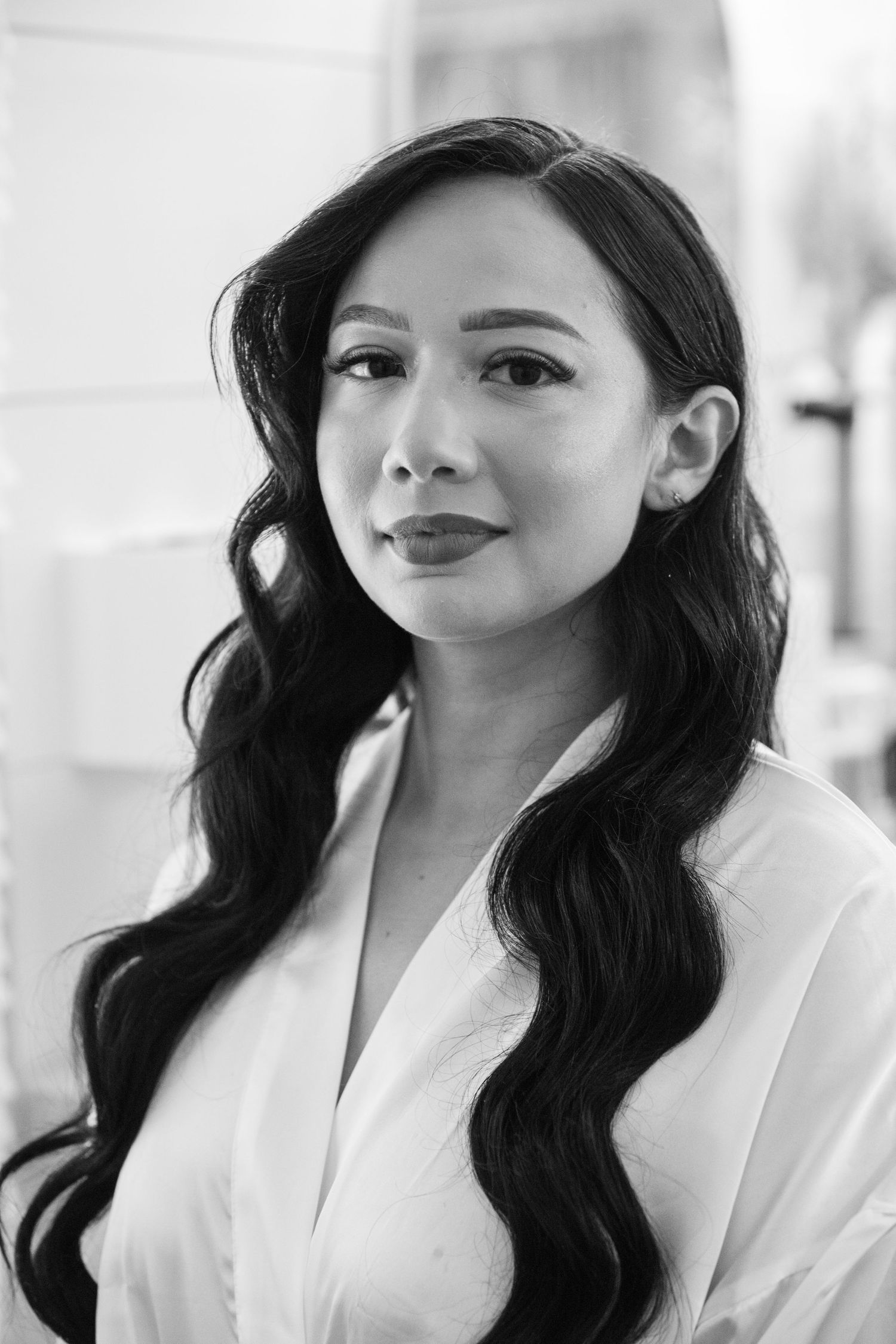 Professional black and white portrait of a woman with long dark wavy hair wearing a white blouse against a light background.
