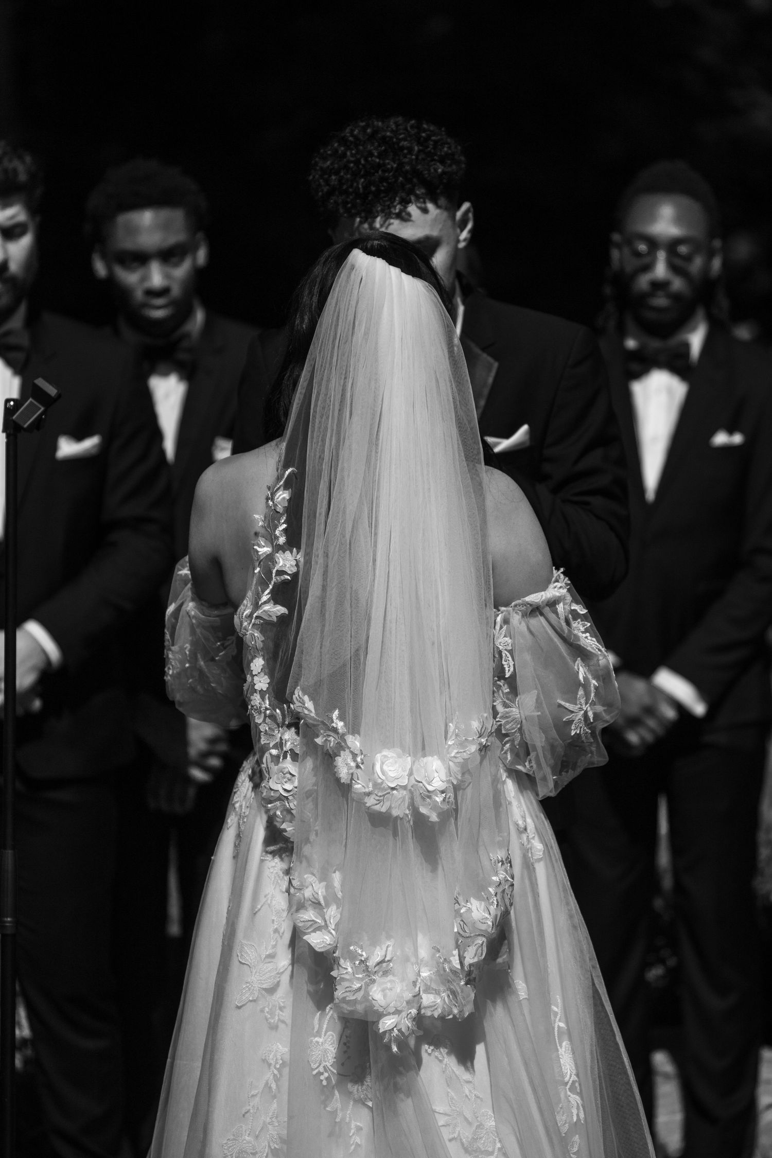 A wedding veil adorned with delicate butterfly appliques is surrounded by guests in formal black attire.