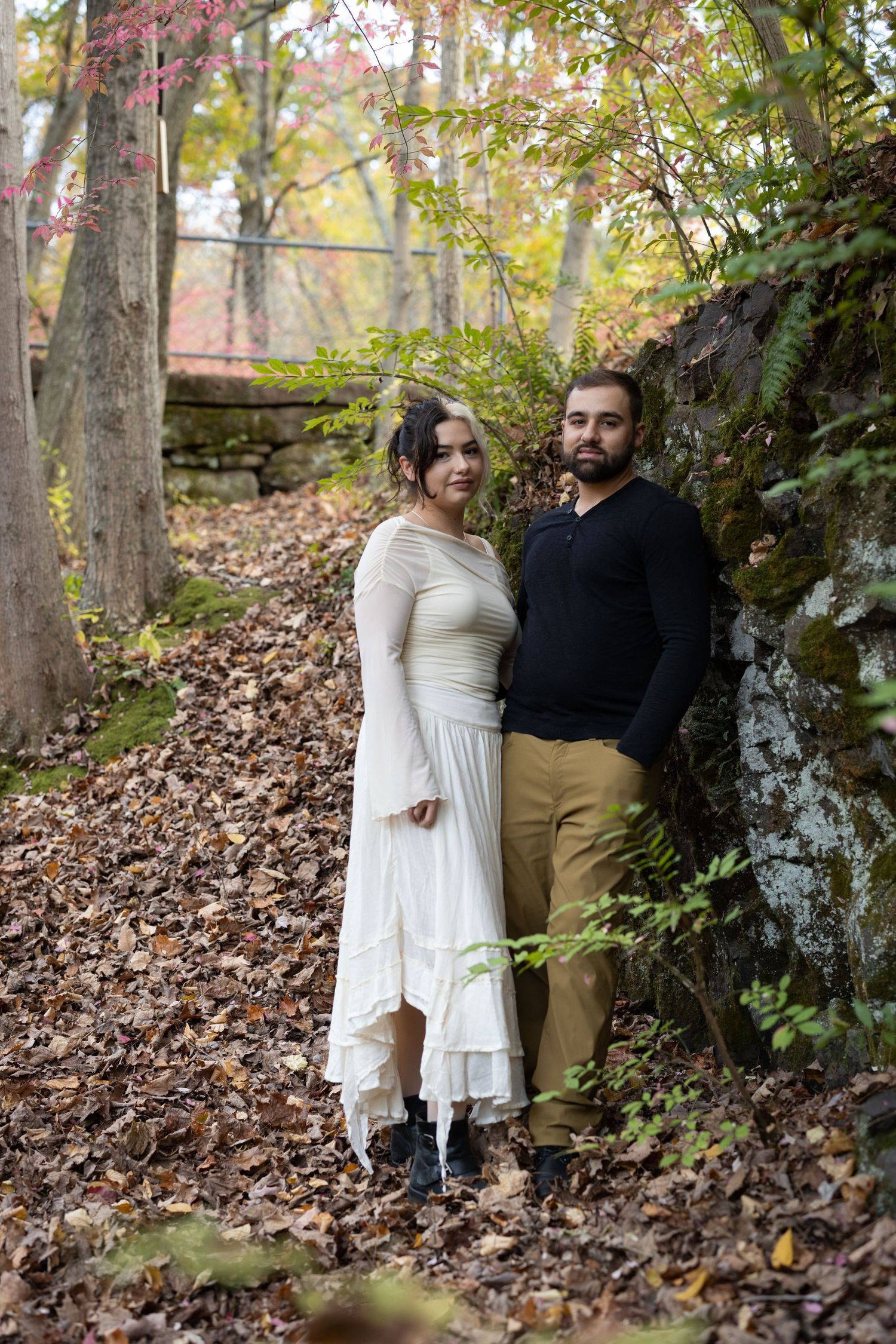Couple posing in autumn forest by mossy rock wall, woman in white dress, man in black sweater and khaki pants.