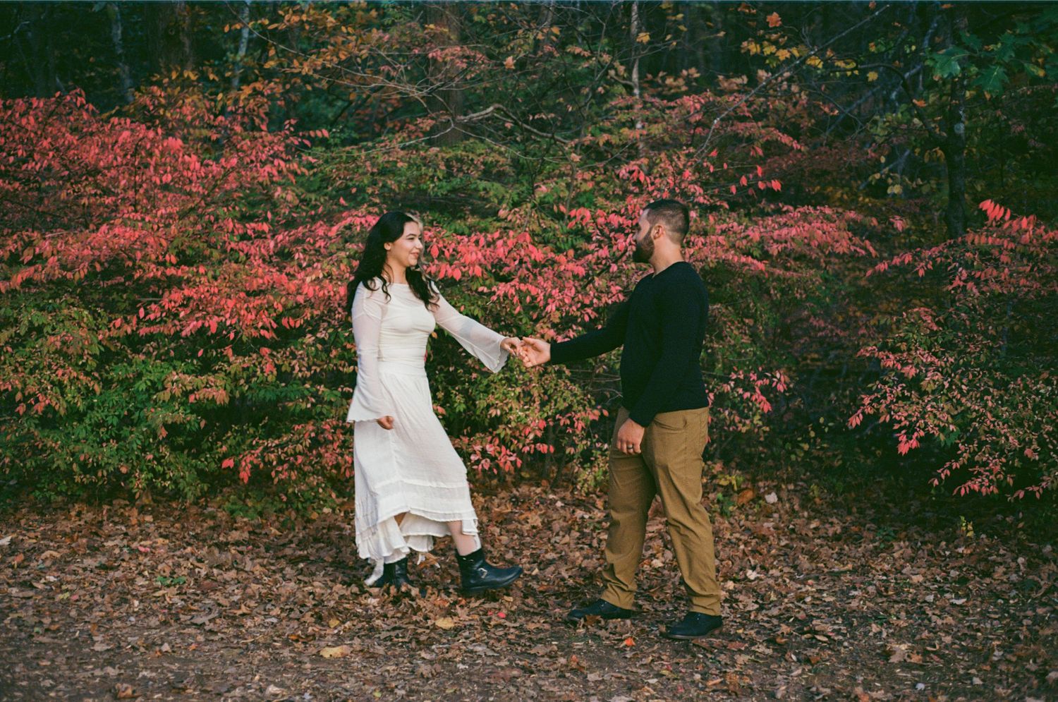 Couple holding hands in autumn forest with vibrant red foliage, woman in white dress and man in black sweater.