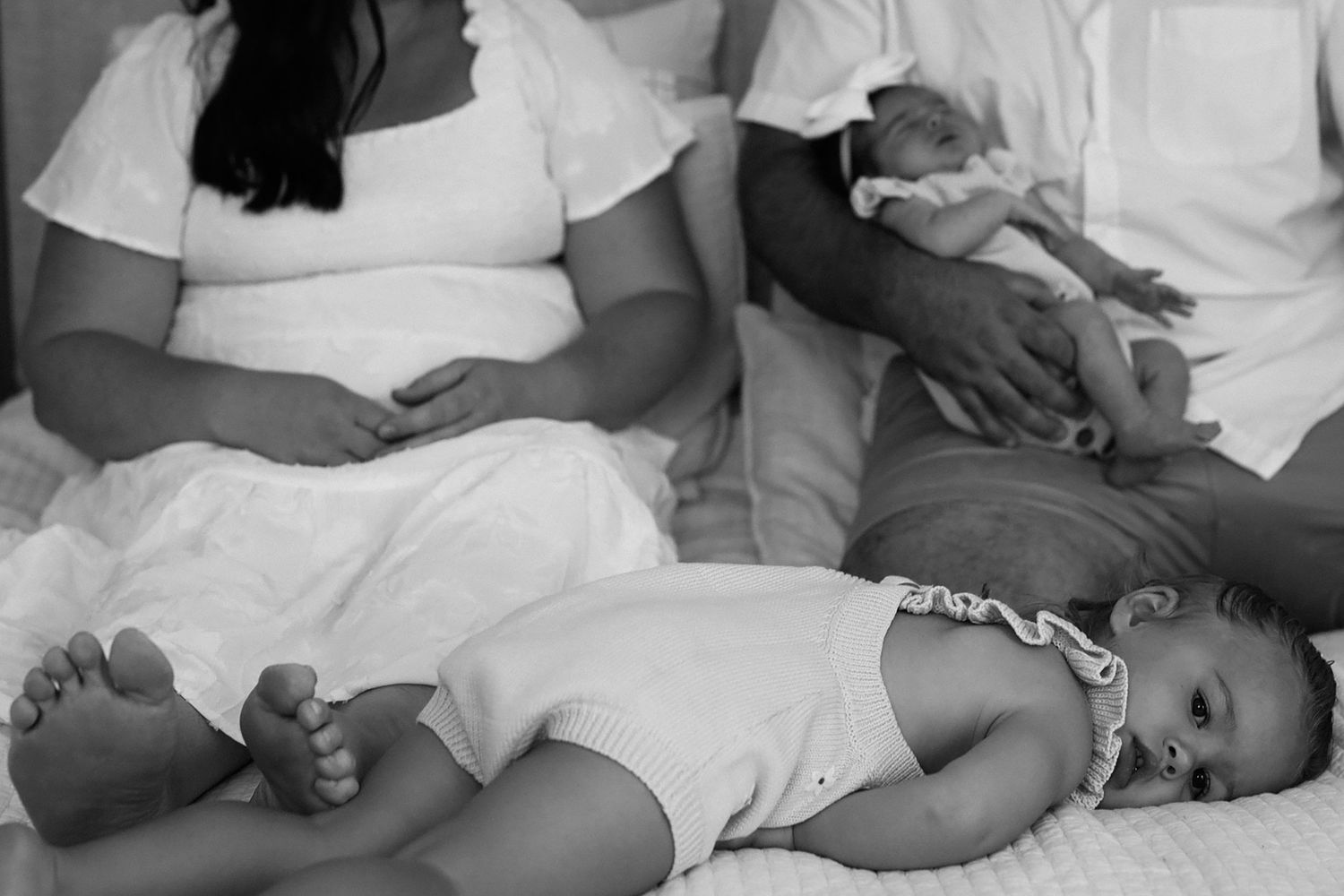 Black and white family portrait of family on bed. Child lays down and looks right at camera. Blank look. Artistic portrait.