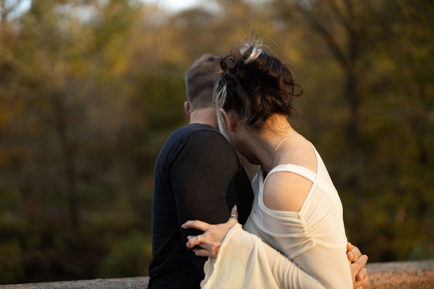 Couple sharing an intimate moment outdoors, woman in white dress embracing man in black shirt with autumn trees in background.