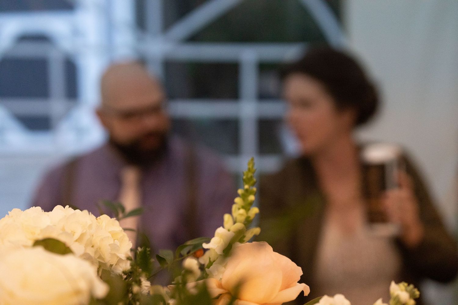 Peach roses and delicate white flowers in a table arrangement with people conversing in the blurred background.