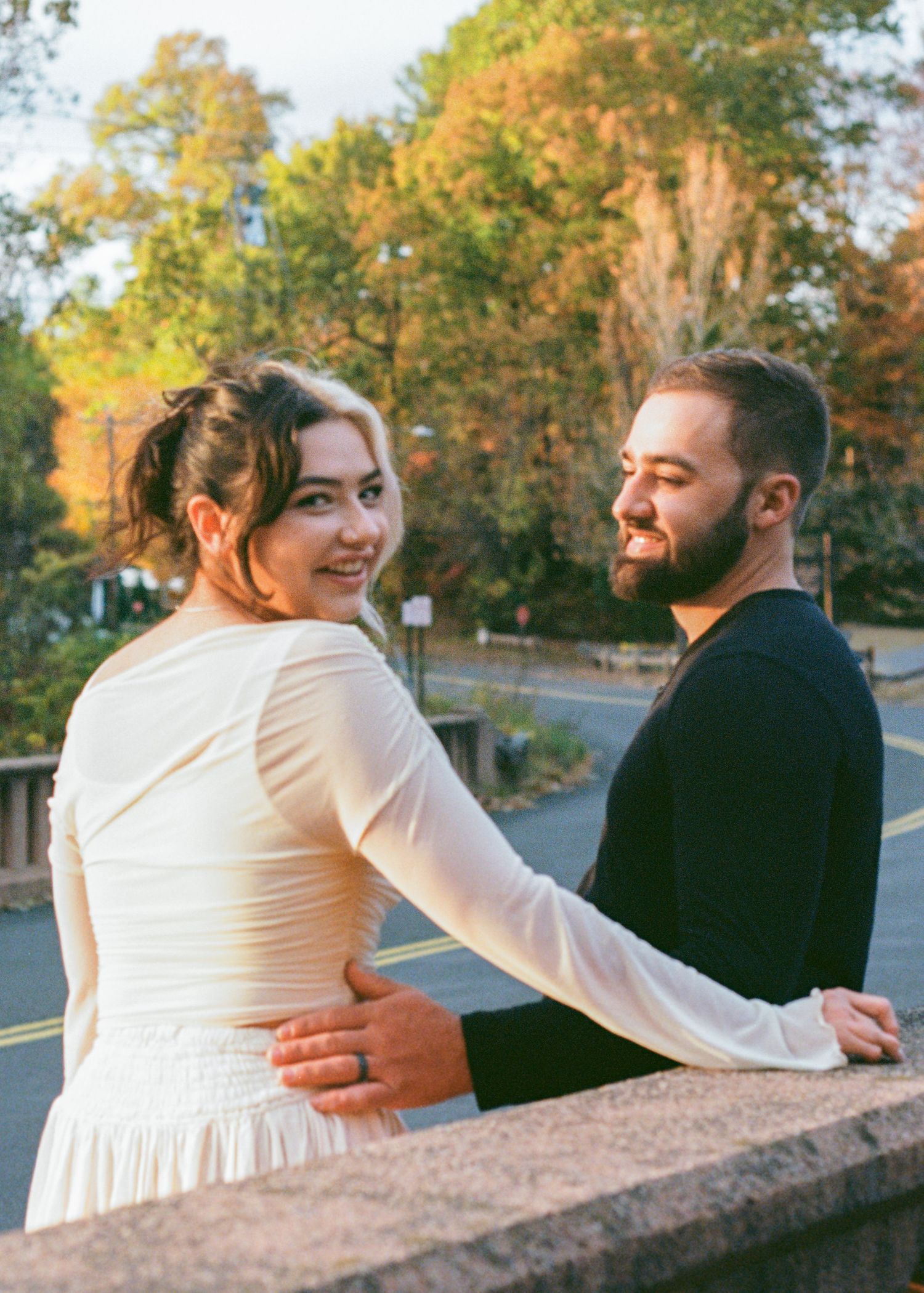 Couple posing on a stone bridge surrounded by autumn foliage, woman in white dress smiling over her shoulder.