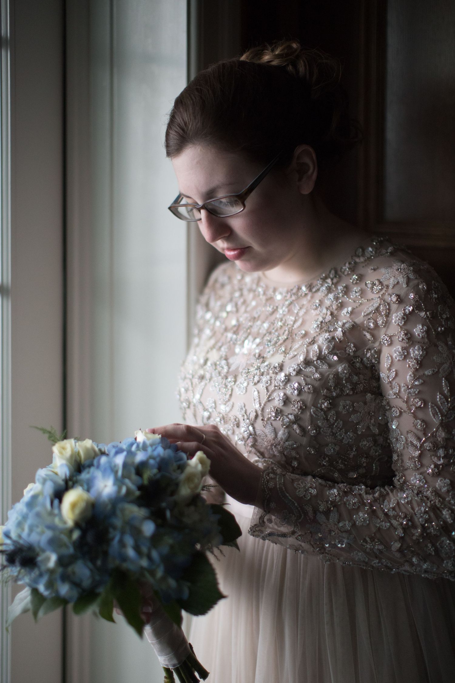 A bride in a lace-sleeved wedding dress gazes at her blue and white bouquet in a series of soft window-lit portraits.
