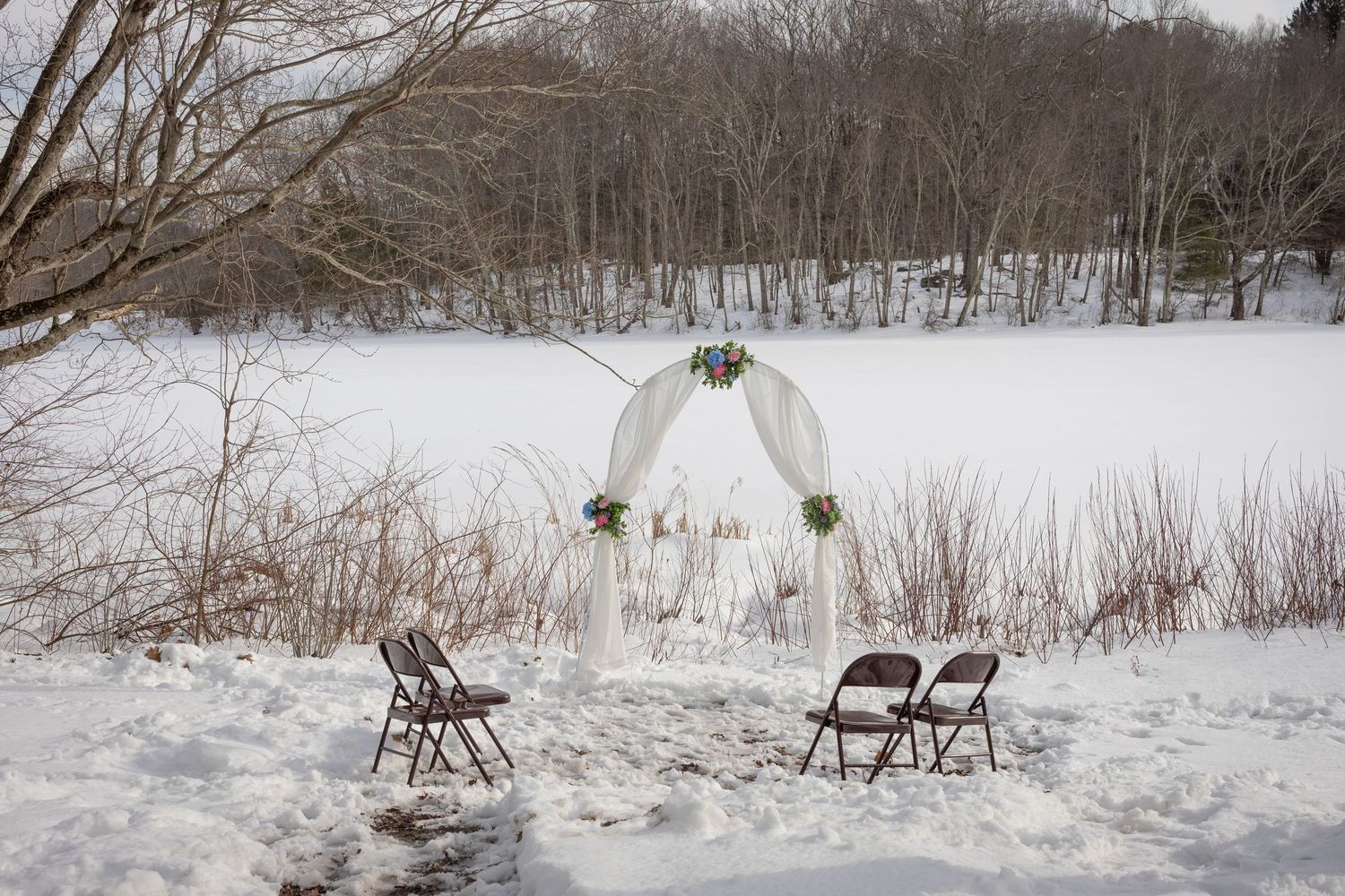 Winter wedding arch draped in white fabric with floral accents, set against a frozen lake and snow-covered landscape.