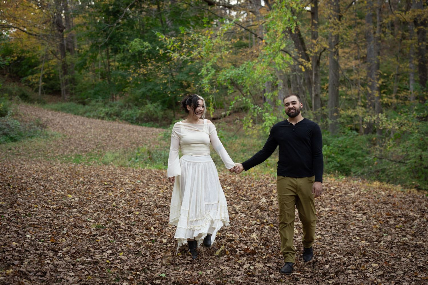 Couple holding hands in autumn forest clearing, woman in white dress, man in black sweater, surrounded by fallen leaves.