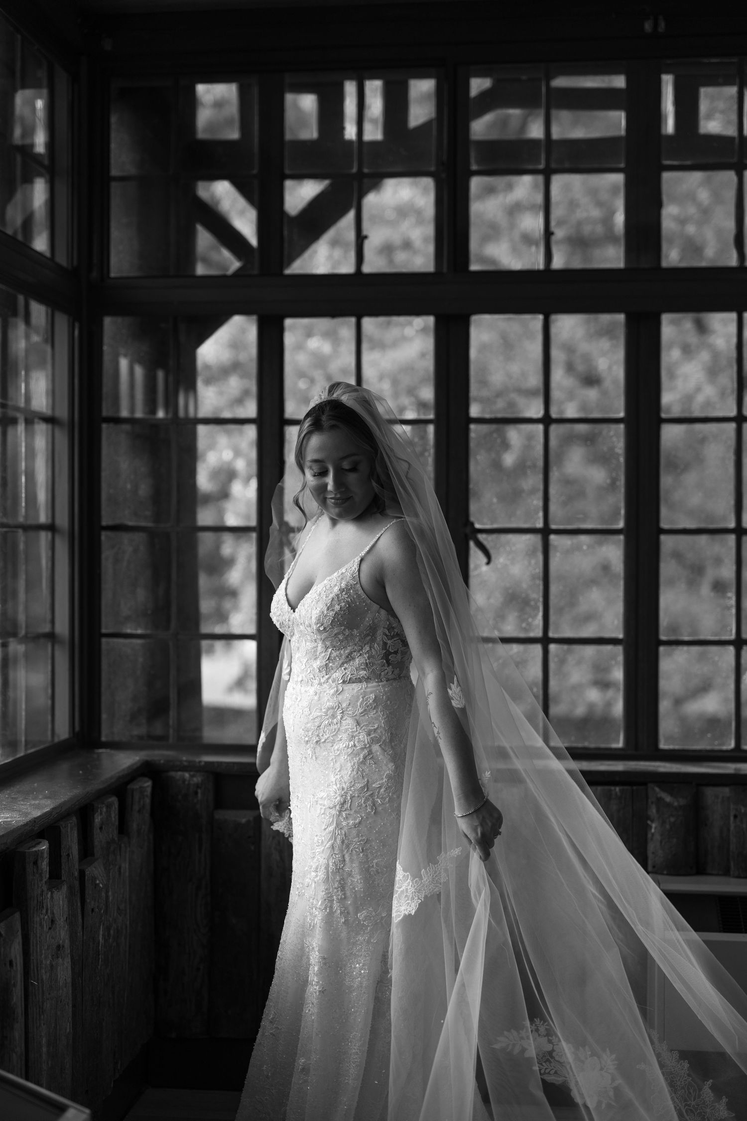 A bride in a white lace wedding dress and veil poses near a window in a rustic wooden building.