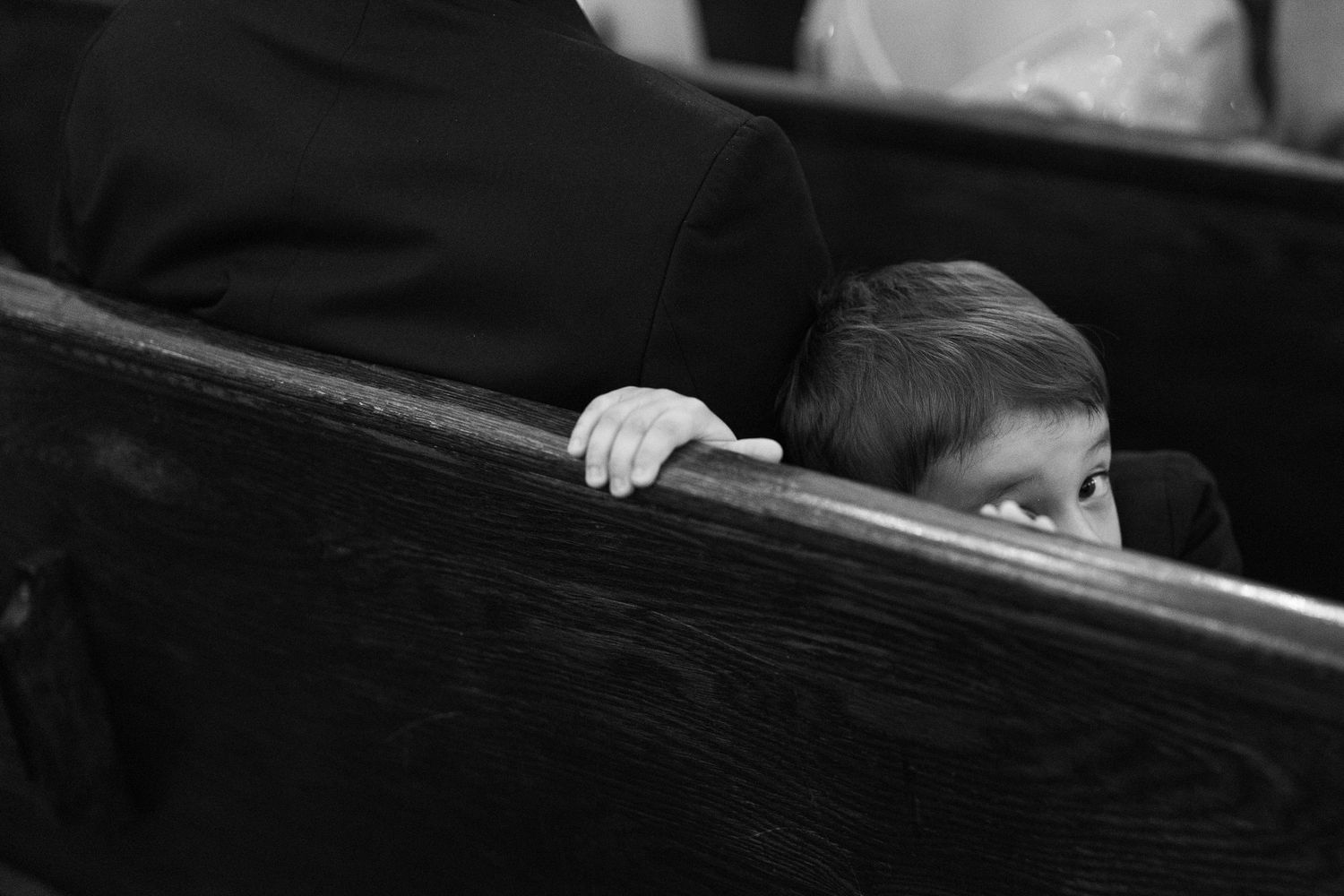 A black and white photograph shows a person sitting in a dark church pew with light streaming through a window. Private ceremony.