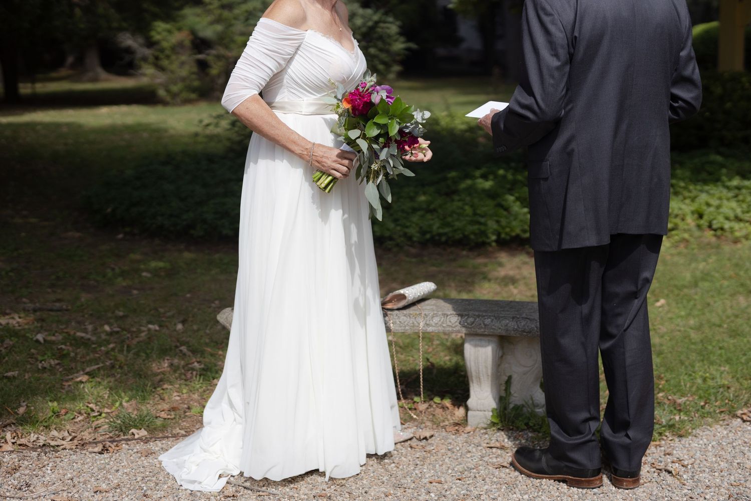 A bride in a white lace wedding dress holds a pink and purple bouquet while standing next to someone in a dark suit outdoors.
