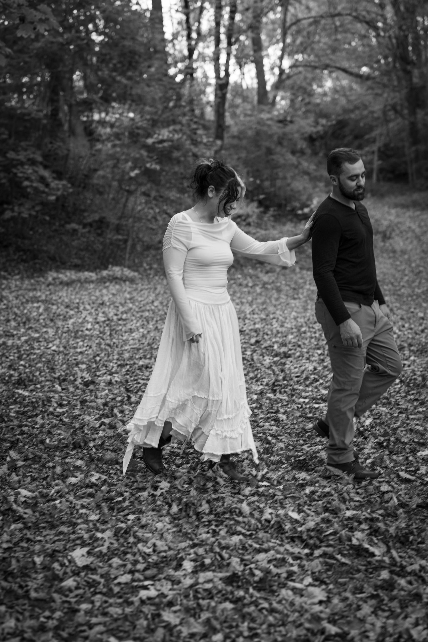 Black and white photo of a couple walking through a leaf-covered forest, woman in white dress, man in dark shirt.