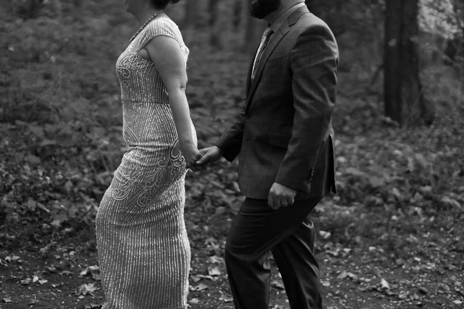 A couple holds hands in a black and white photo while standing in a wooded forest setting.