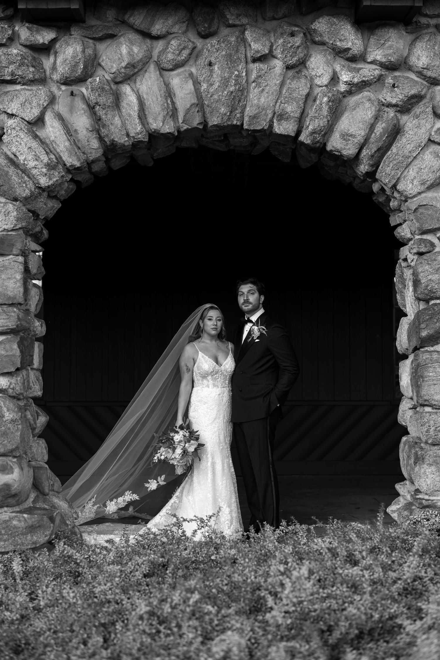 Black and white wedding portrait of bride and groom standing in a stone arch doorway, bride holding bouquet with veil flowing.