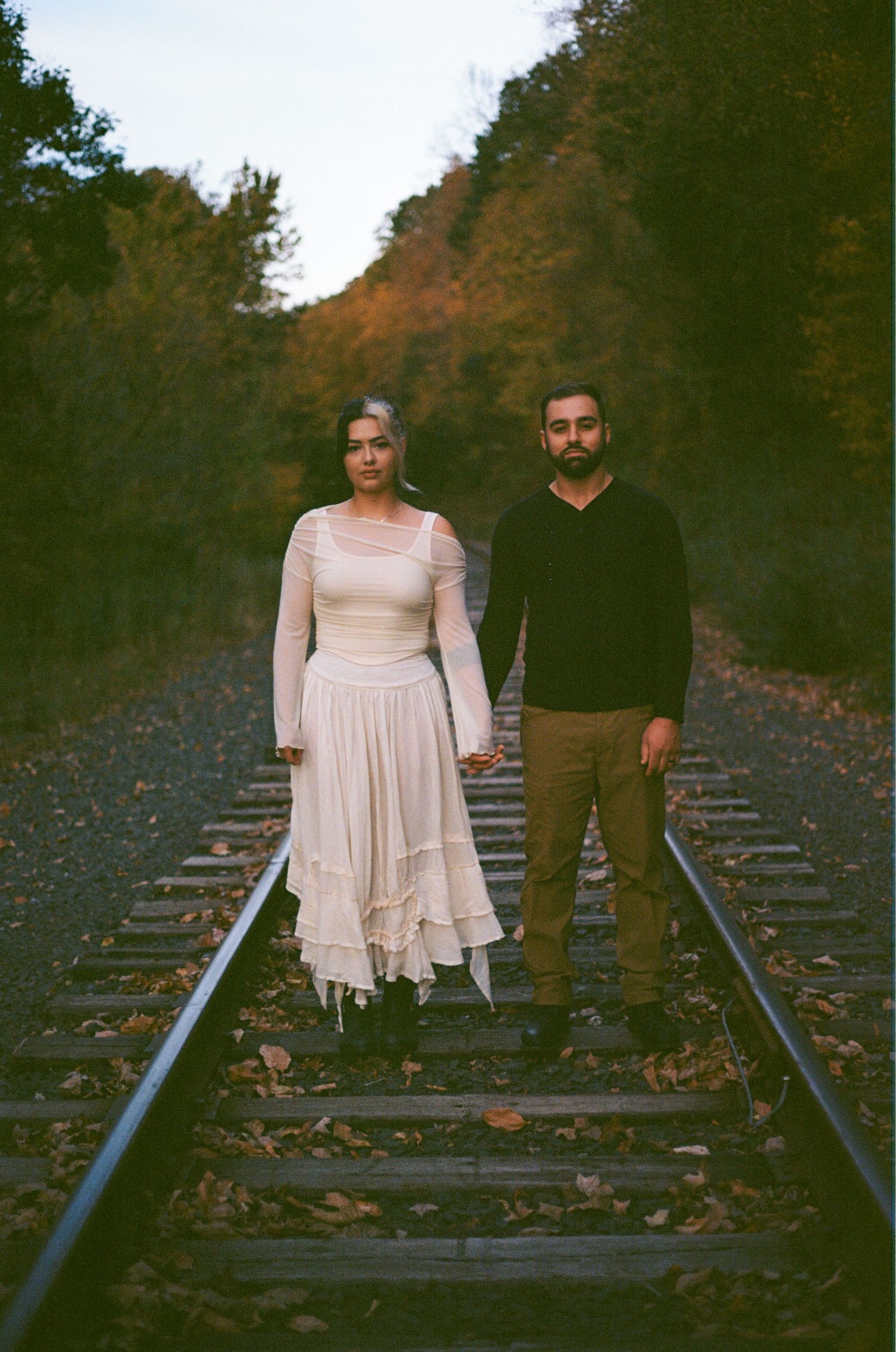 Couple holding hands on railroad tracks surrounded by autumn foliage, woman in white dress, man in black sweater.