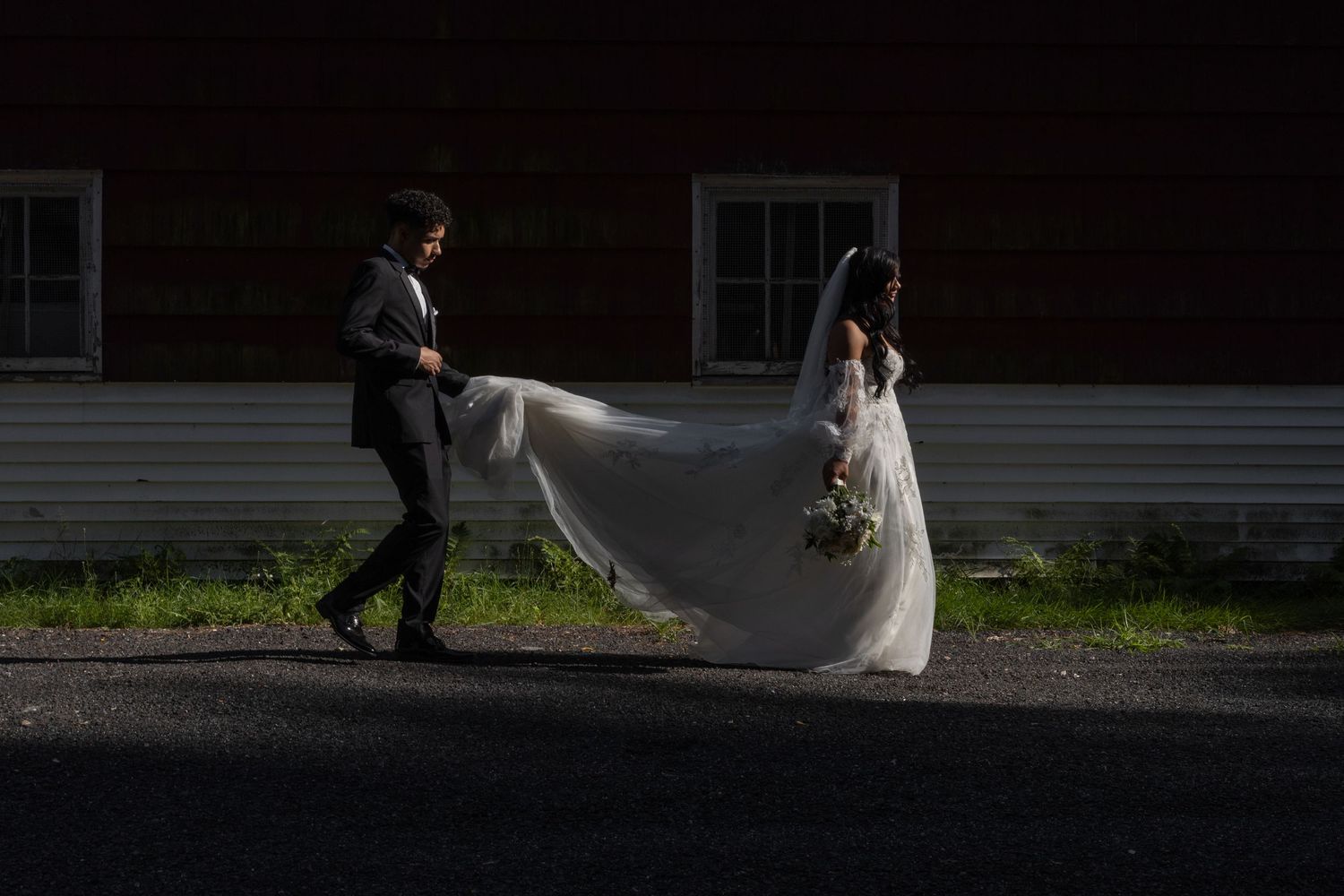 A dramatic wedding photo with the bride's veil flowing in the light against a dark rustic building backdrop.