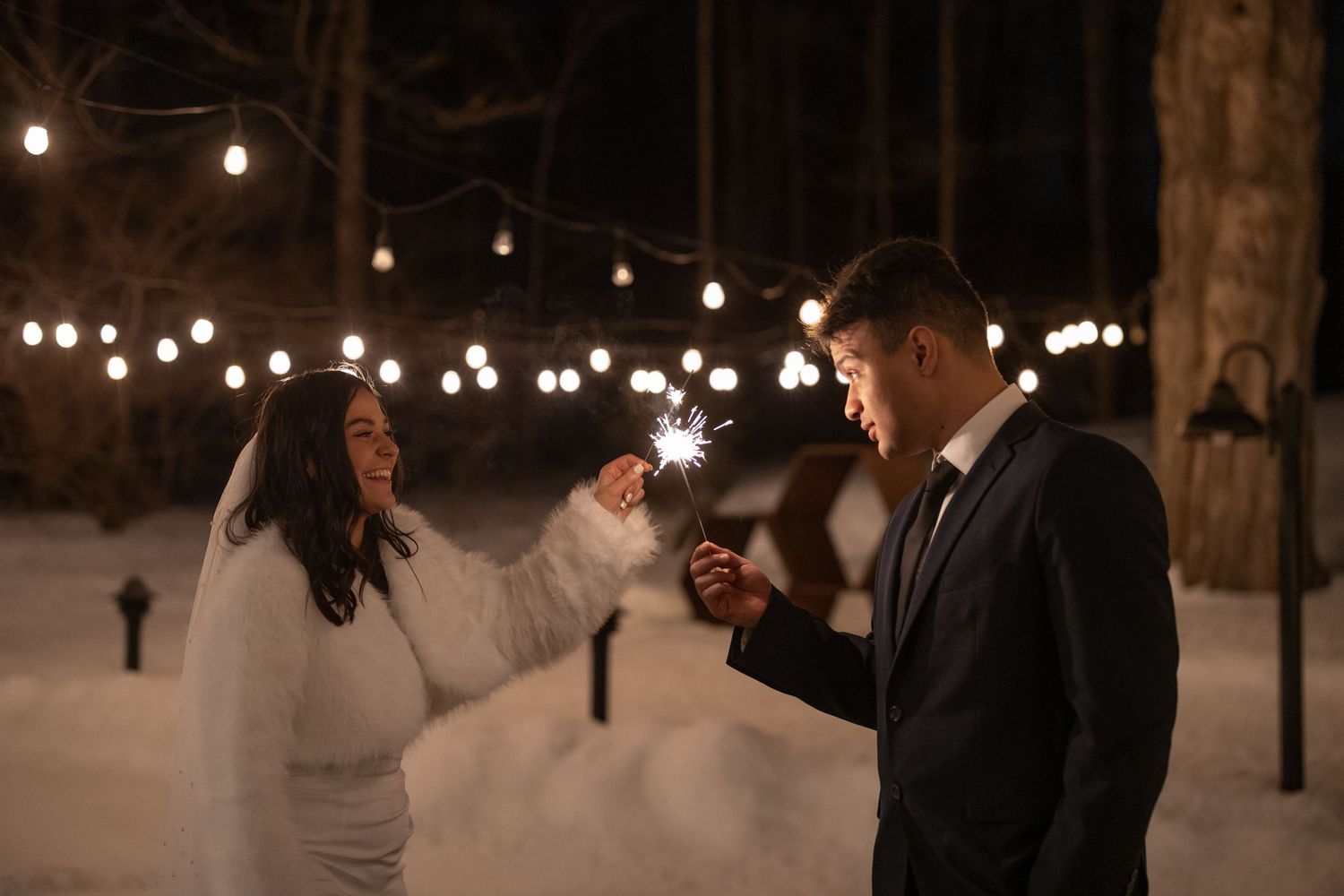 Bride and groom holding sparklers outdoors at night in winter, with string lights glowing in snowy forest background.