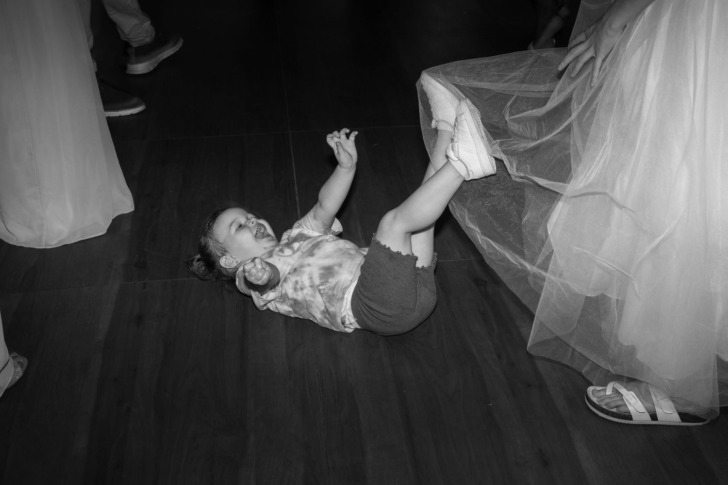 A young child lays on a dance floor laughing and playing with a flowing white veil at wedding reception.