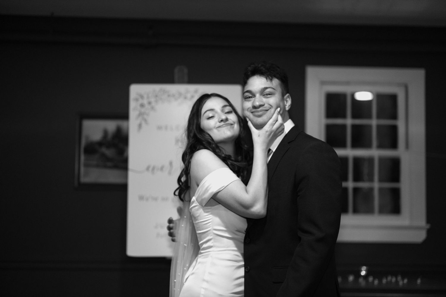 Black and white photo of a couple embracing and smiling at a formal event, woman in white dress hugging man in suit.