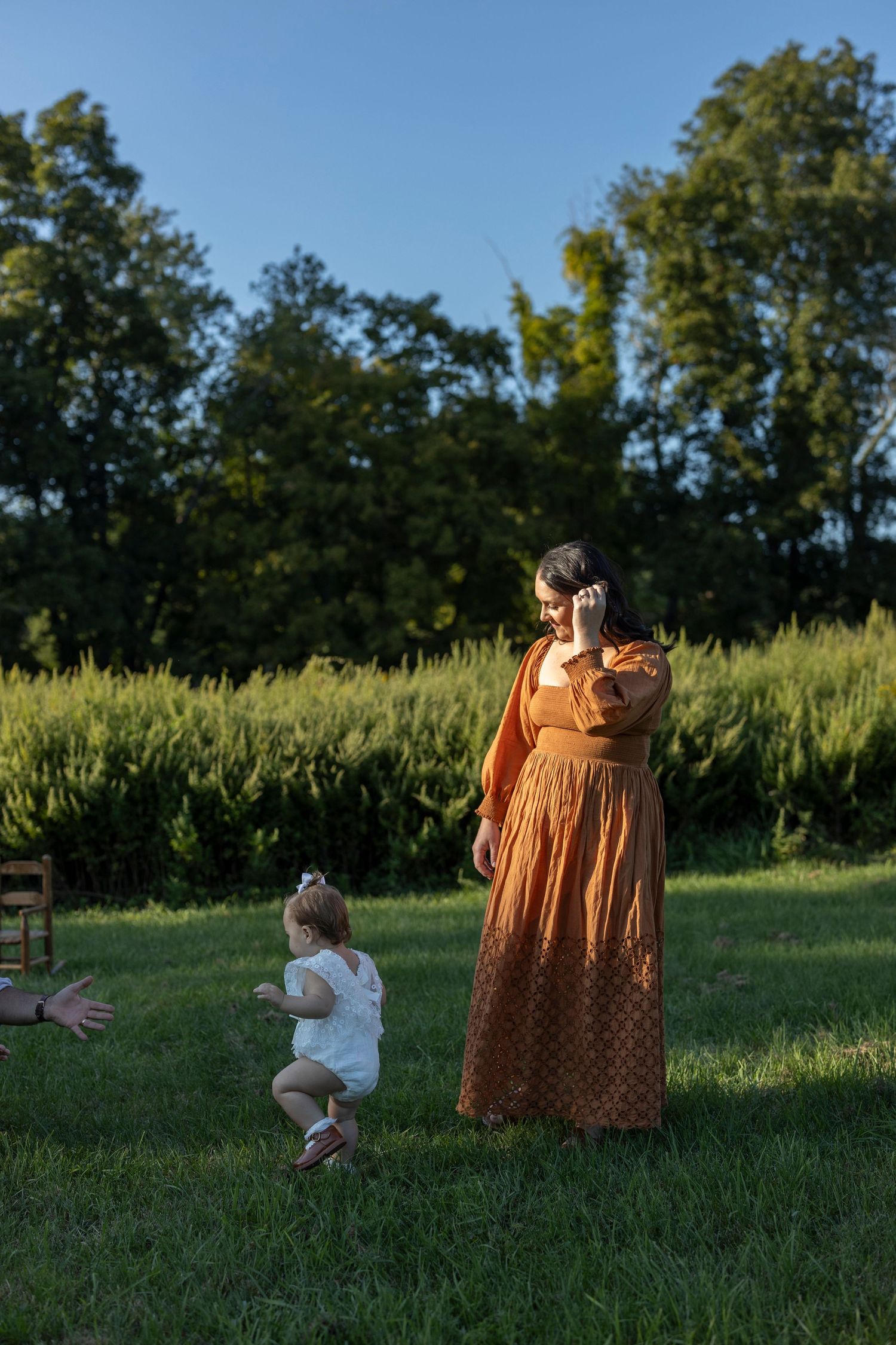 Mother standing next to her child in warm direct sunlight as her baby walks towards her dad.