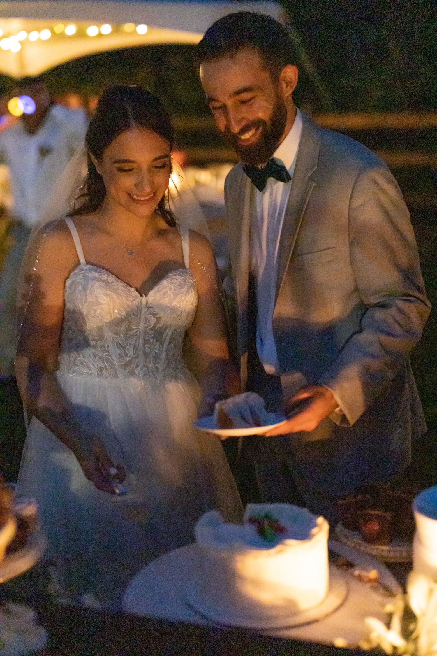 A joyful couple cuts their wedding cake together at an evening reception with twinkling lights in the background.
