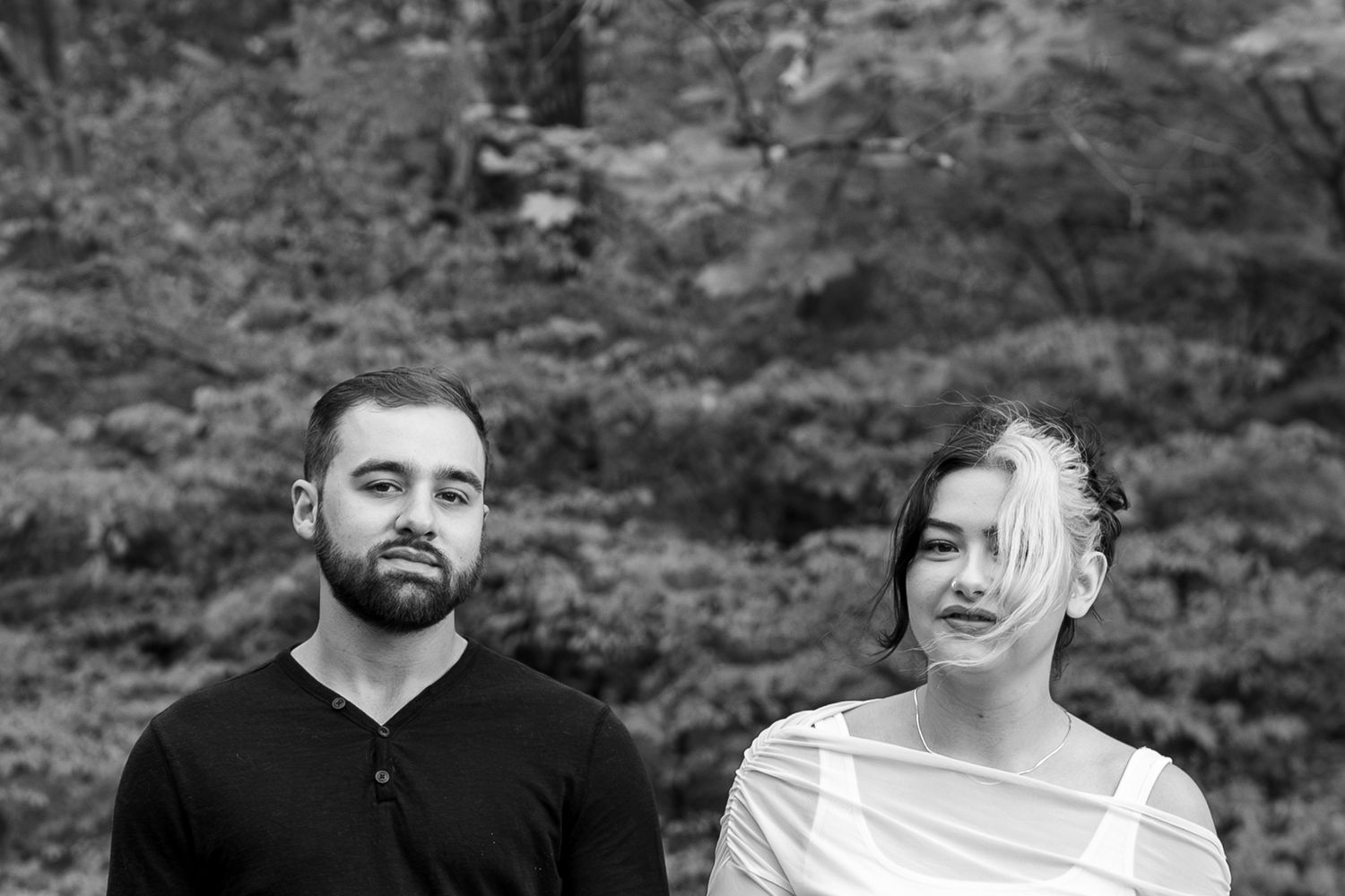 Black and white portrait of a bearded man and young woman standing outdoors with lush trees in the background.