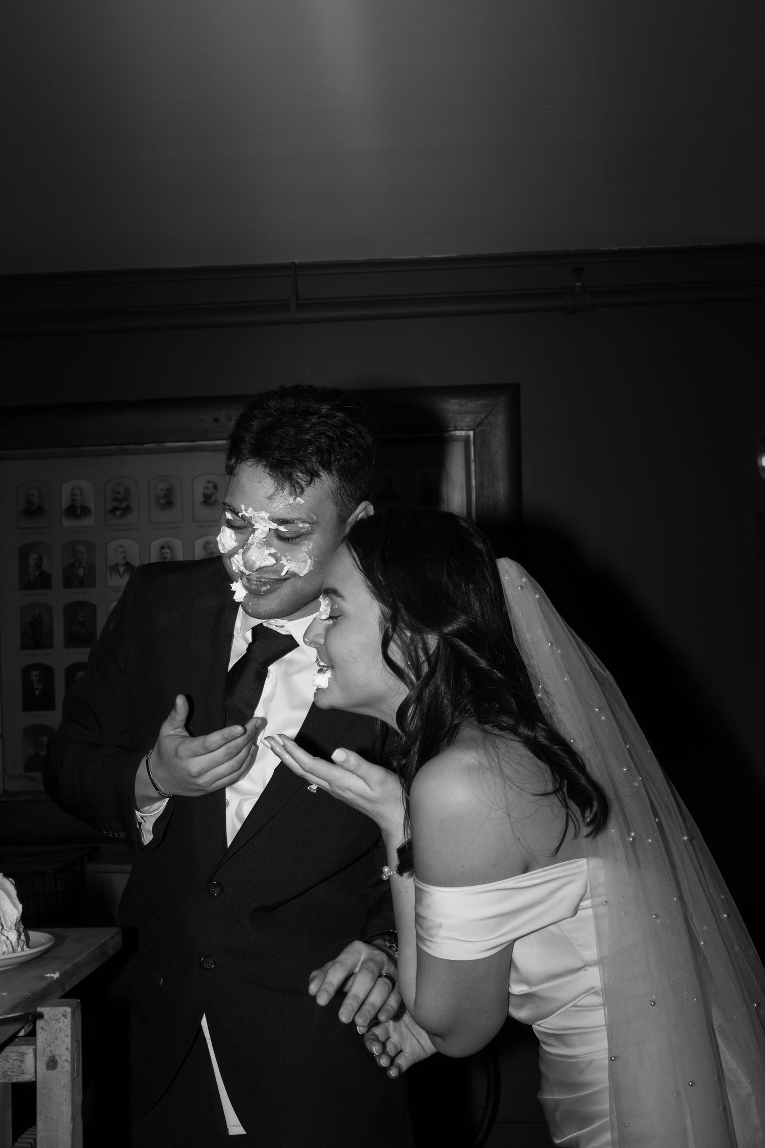 Black and white photo of bride and groom laughing with cake on their faces during wedding reception cake cutting.