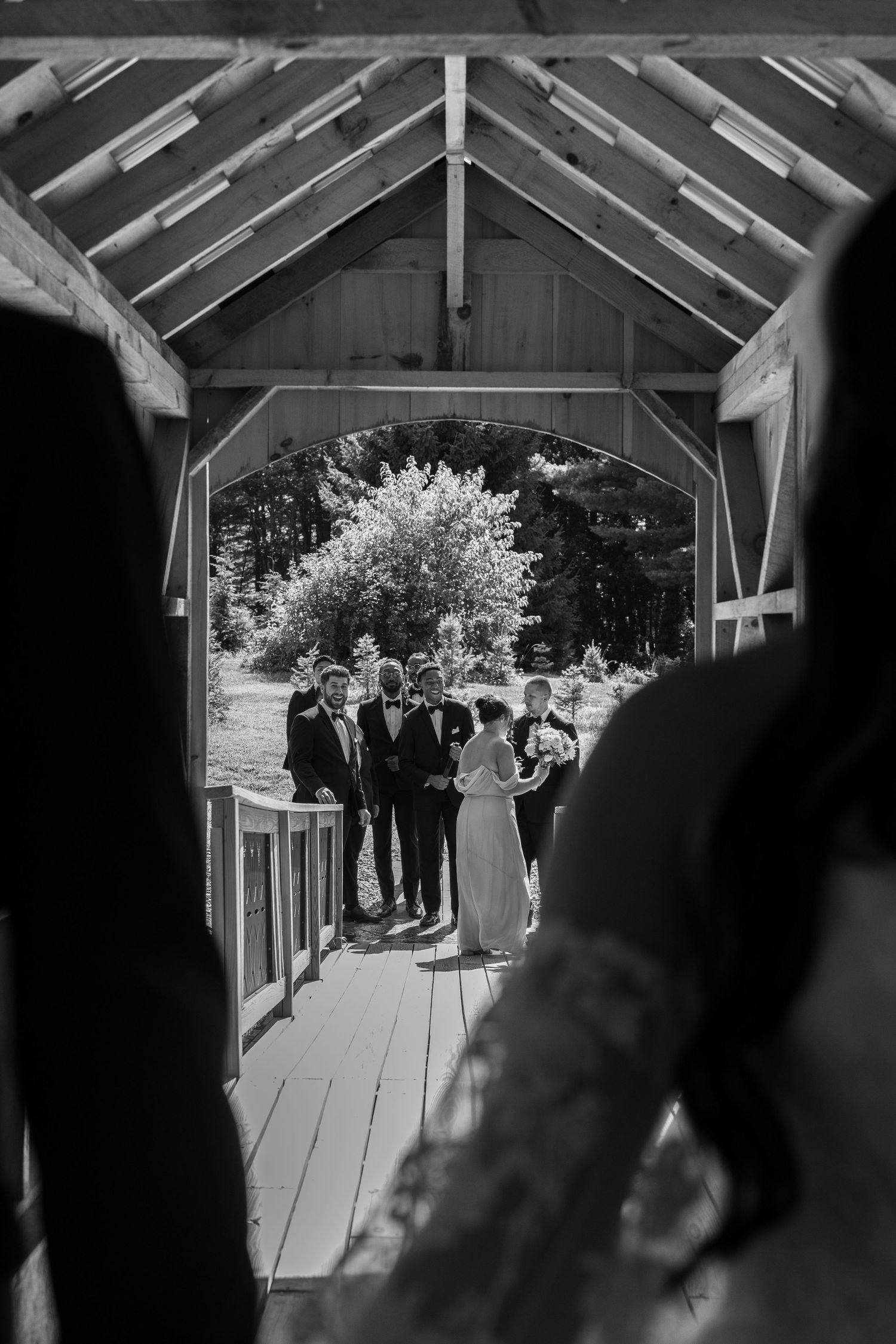 Black and white portrait of a bride and groom standing on a covered Bridge while their bridal party watches.