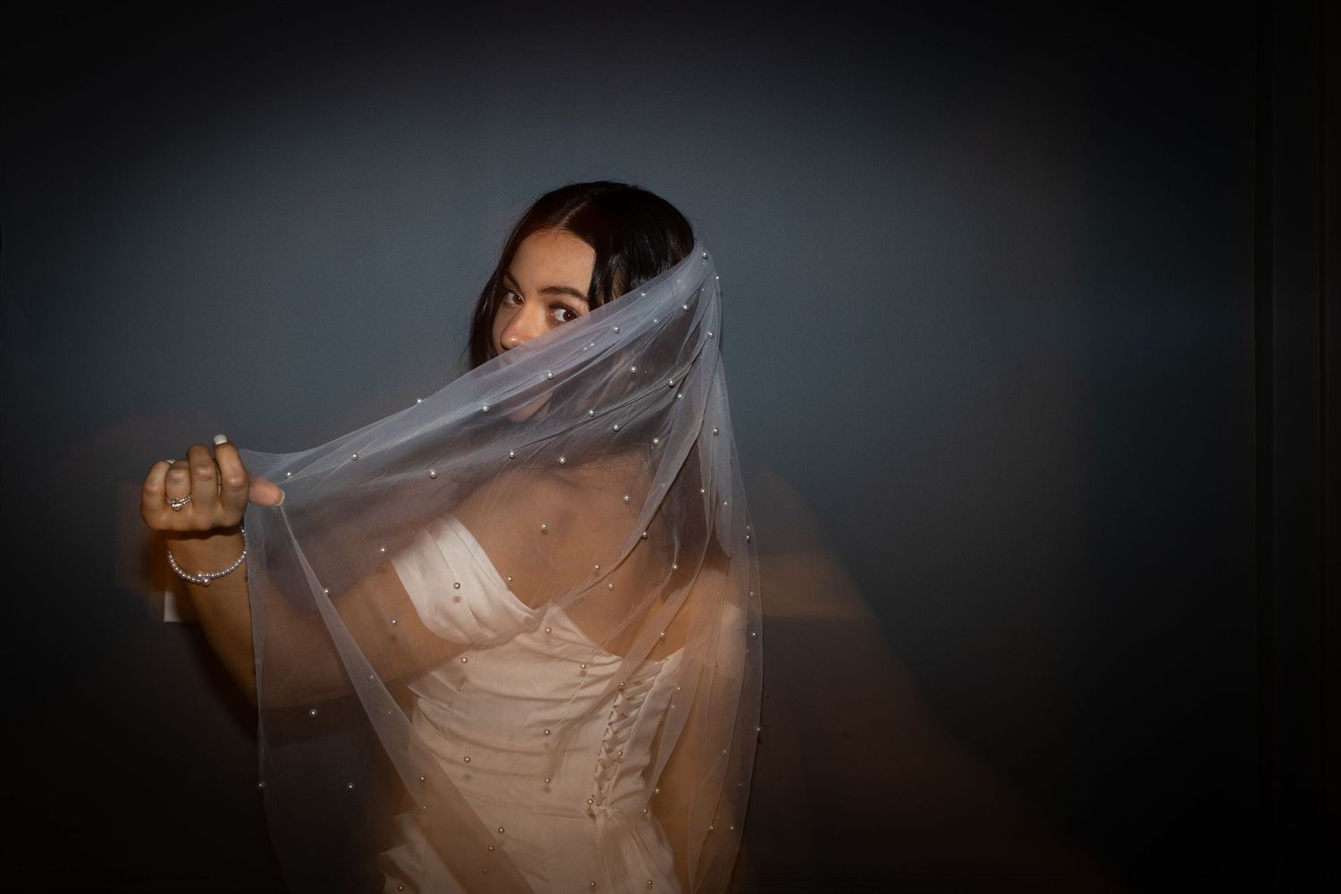 Bride playfully holding her pearl-embellished wedding veil over her face in a dramatic dark studio portrait.
