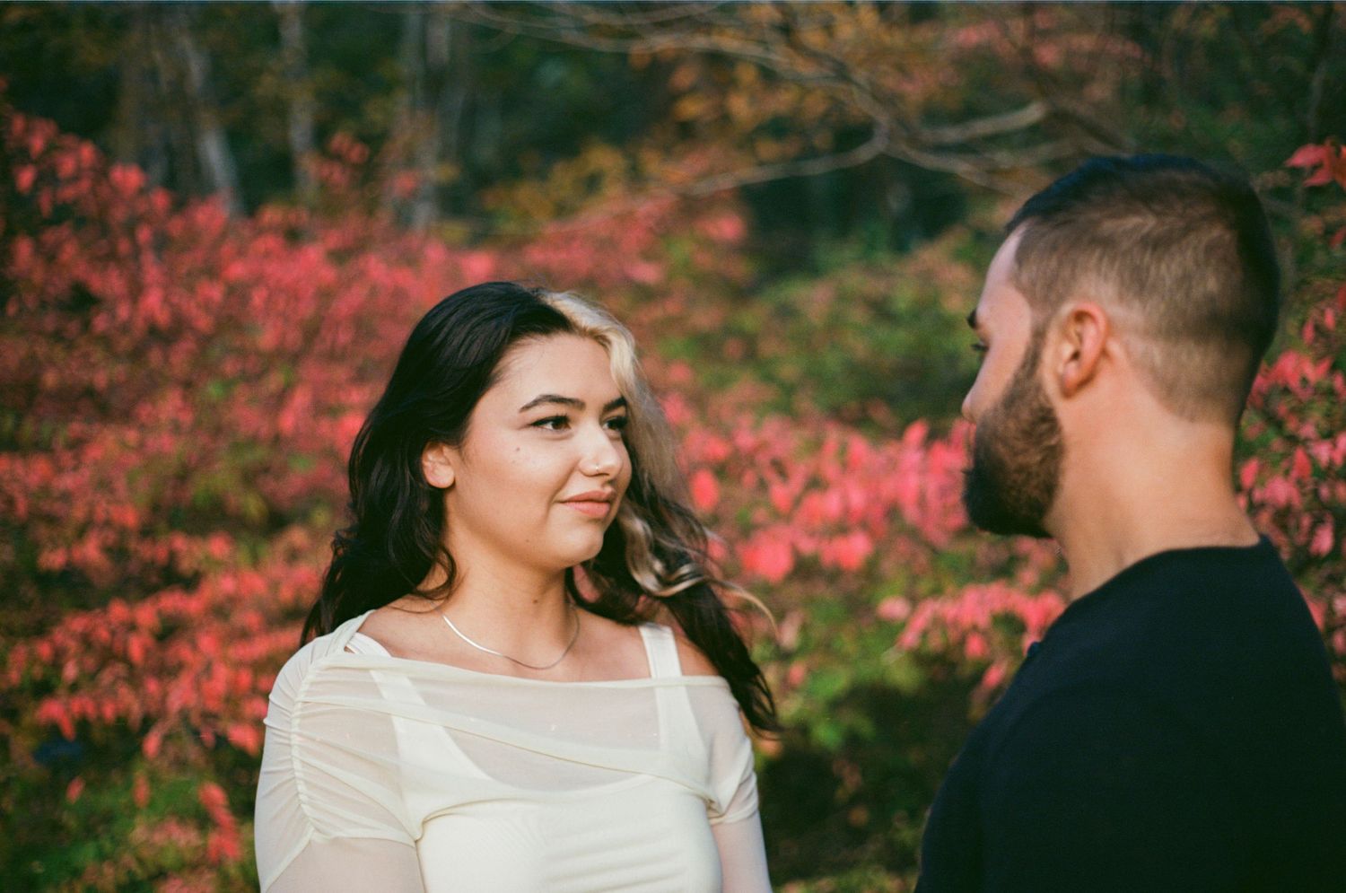 A couple shares a tender moment outdoors surrounded by vibrant red autumn foliage during an engagement photo session.