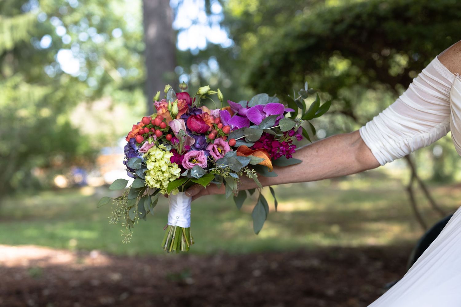 A bride in a white wedding dress holds a vibrant bouquet of purple and yellow flowers in an outdoor garden setting.