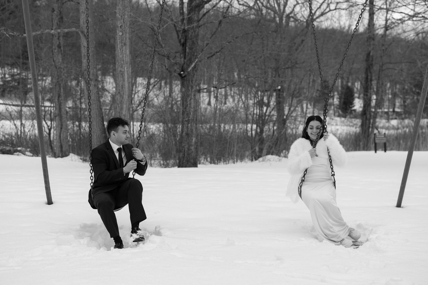 Black and white winter wedding photo of bride and groom playing in snow among bare trees.