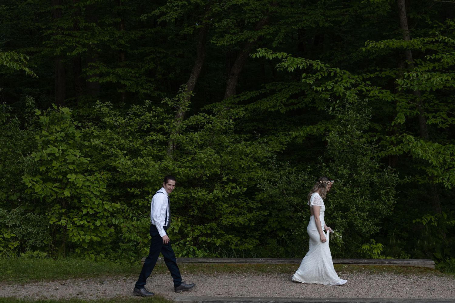 Two people in formal wedding attire walk separately along a path with lush green forest in the background.