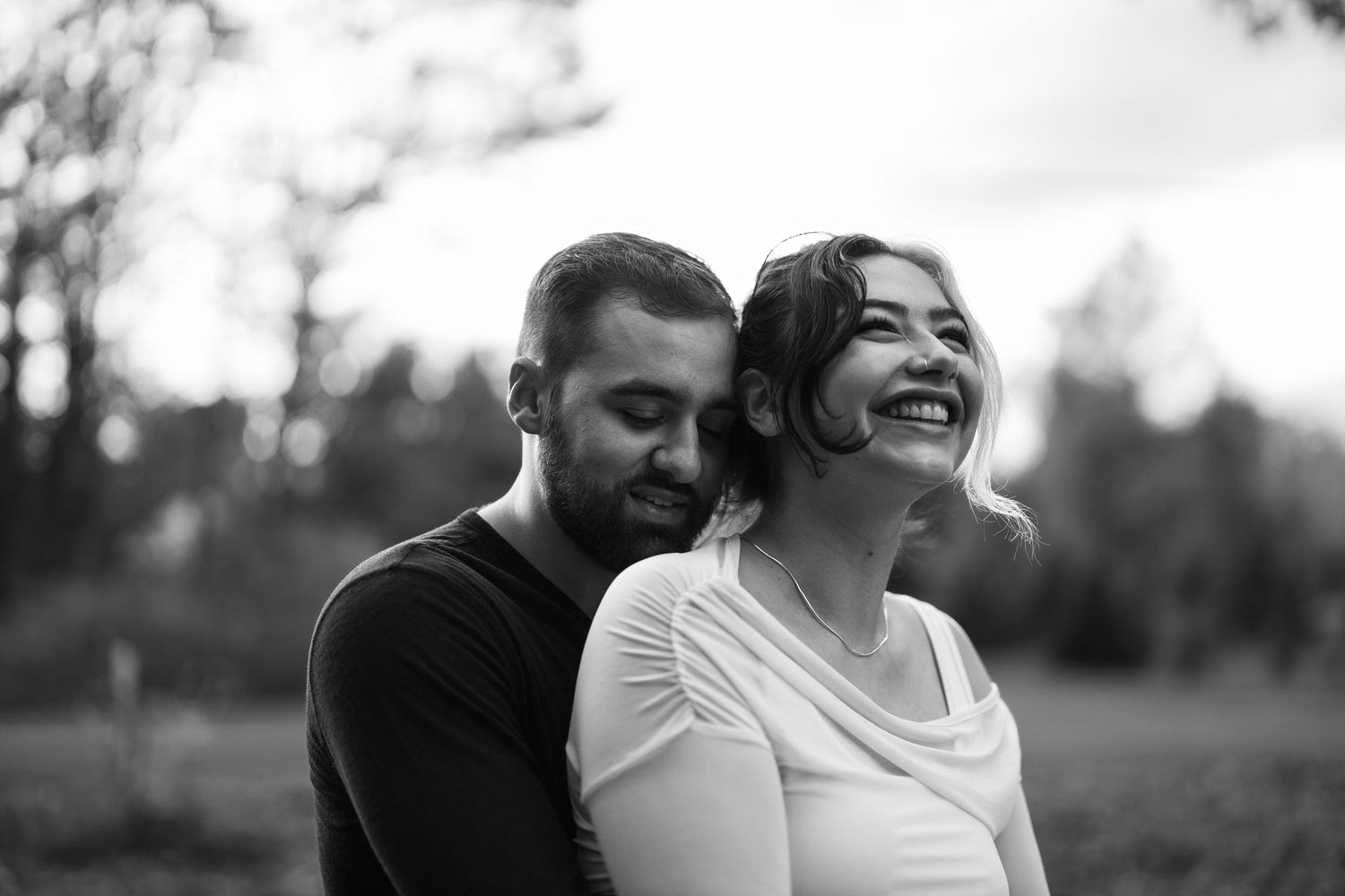 Black and white photo of a couple embracing outdoors, woman laughing joyfully as man nuzzles her neck.