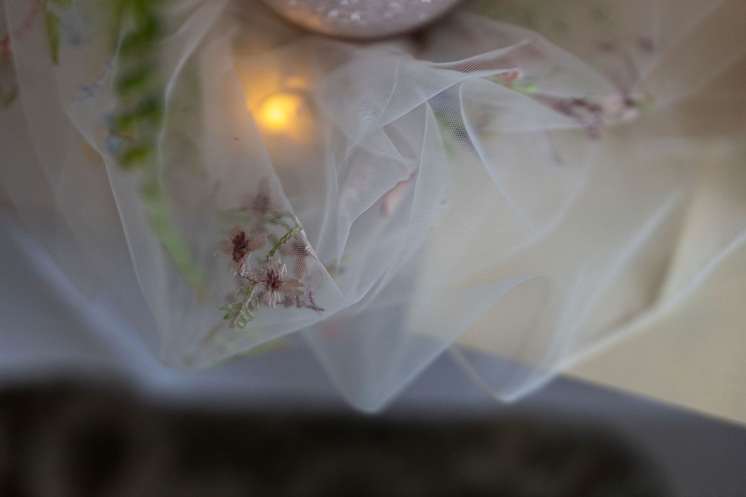 Sheer floral fabric with delicate pink flowers draped softly over a glowing candle in a dreamy, ethereal setting.