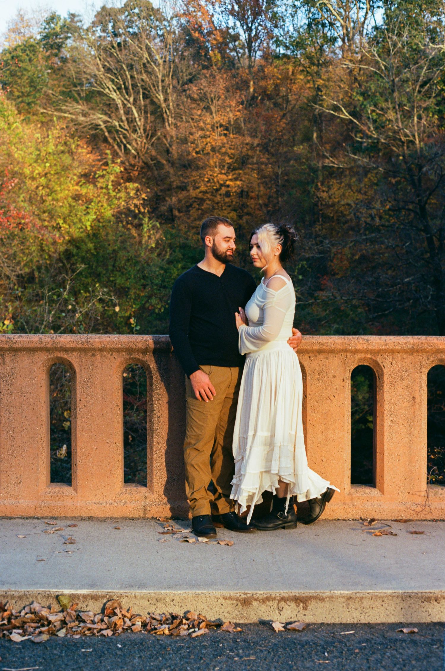 Couple embracing on a stone bridge surrounded by colorful autumn foliage, woman in white dress and man in black sweater.