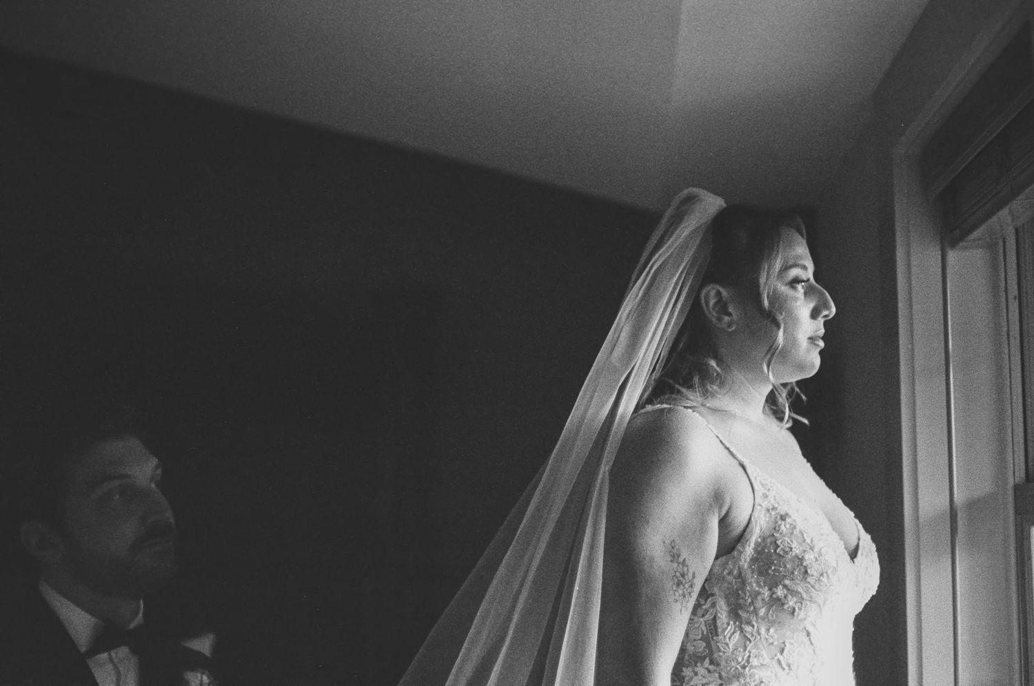 Black and white wedding portrait shot on 35mm film of bride in lace gown and veil gazing out window with groom in tuxedo behind her.