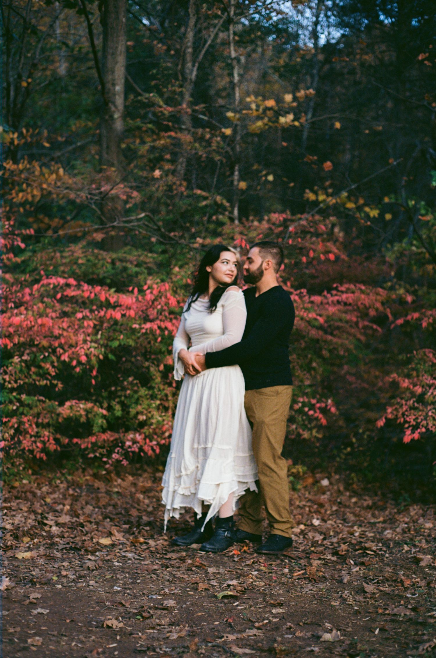 A romantic couple shares tender moments together among autumn foliage and pink flowering bushes in a wooded setting.