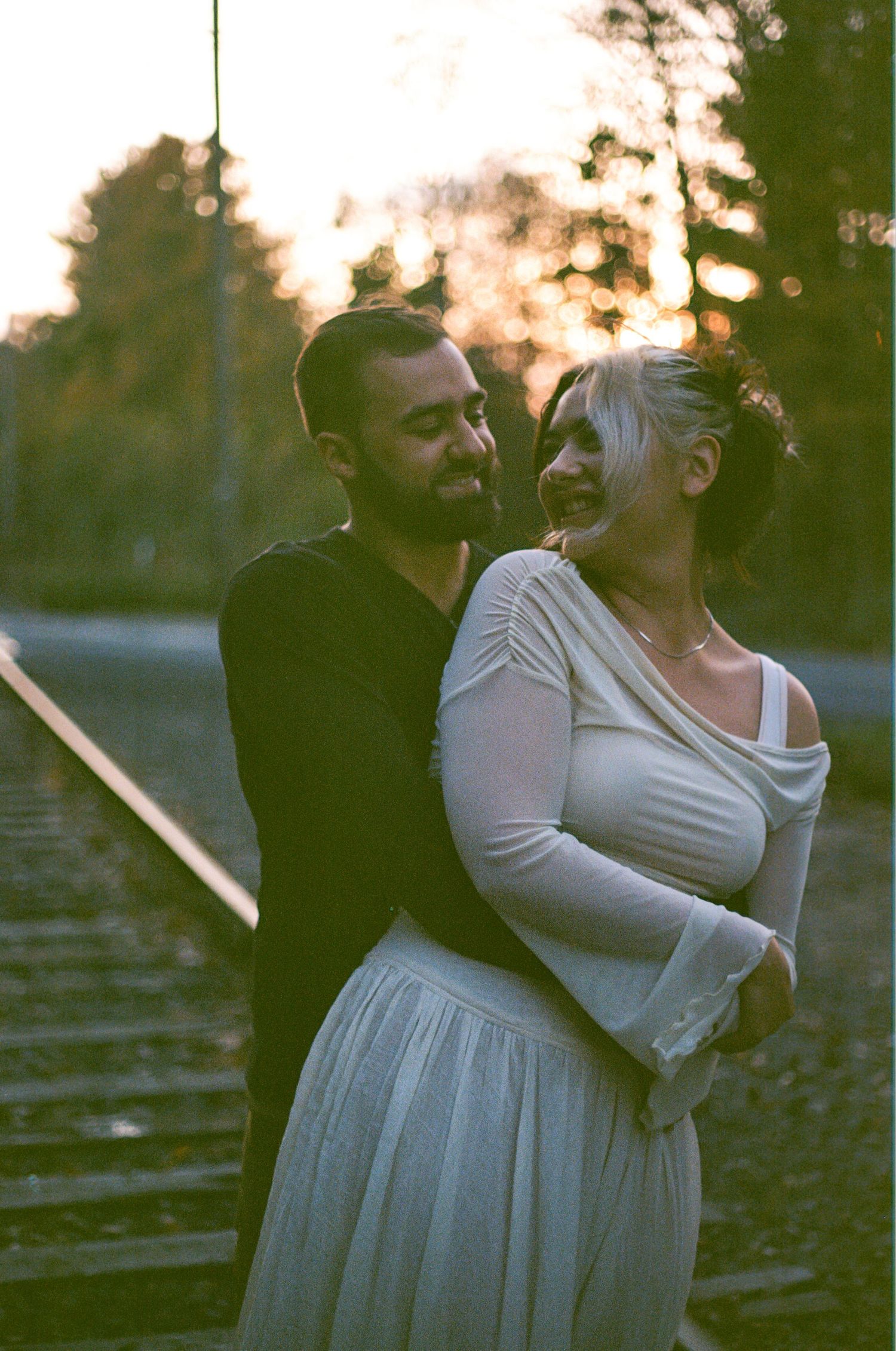 A couple embraces on railroad tracks at sunset creating a romantic silhouette against the golden sky. 35mm film portrait.