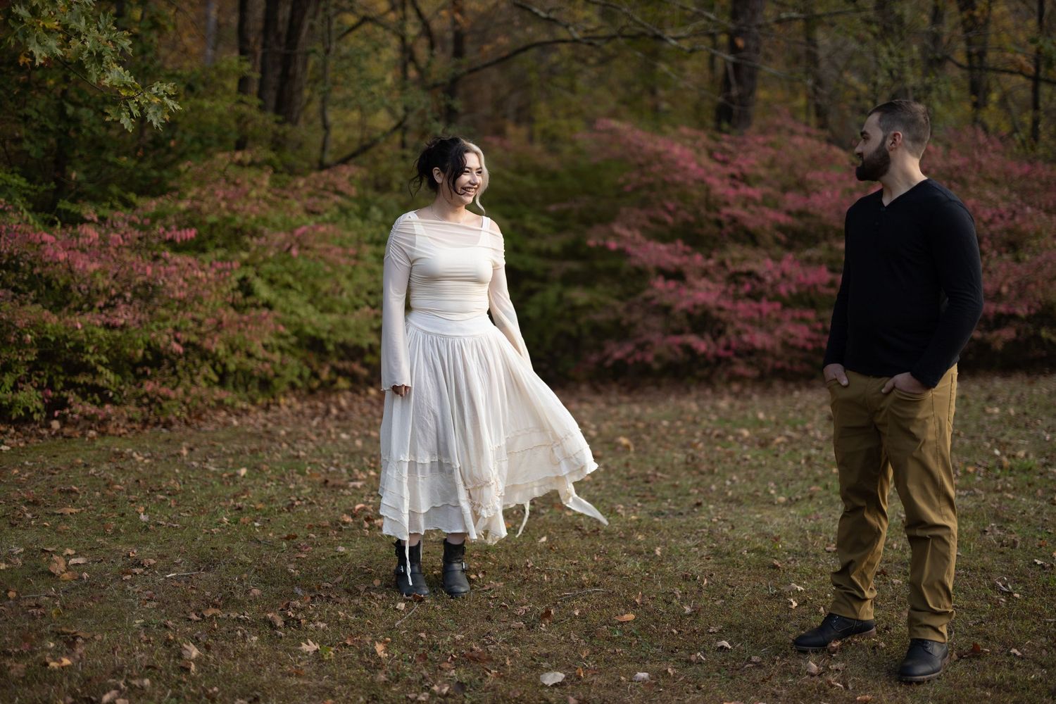 Couple standing outdoors in autumn forest, woman in white dress smiling at bearded man in black sweater and khaki pants.