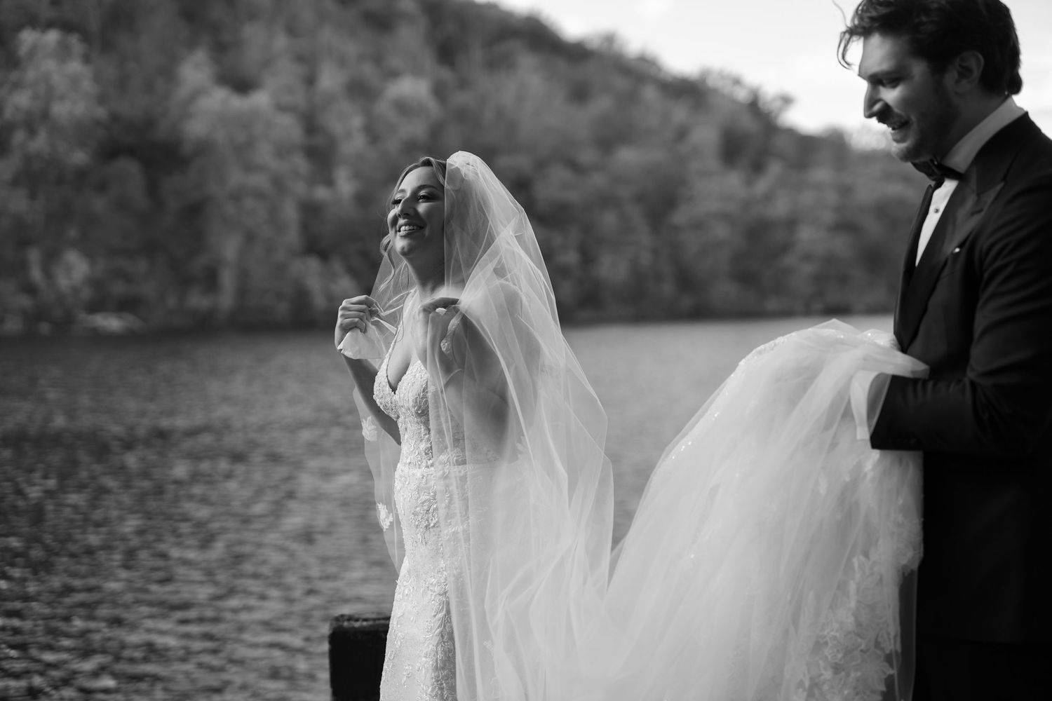 Black and white wedding photos showing couple in romantic poses by a lake with flowing veil and formal attire.