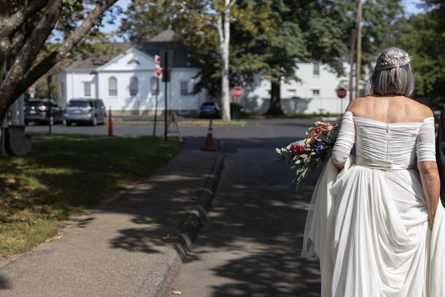 A bride in a flowing white dress walks along a tree-lined path on a sunny day.