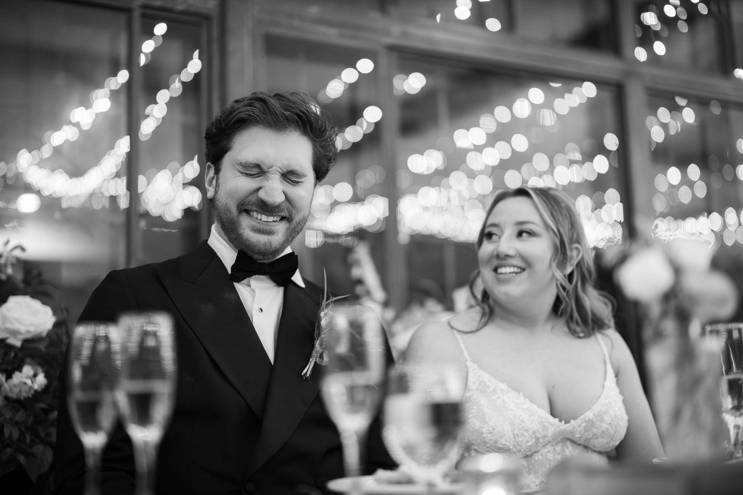 A bride and groom share a joyful moment together at their wedding reception under twinkling string lights.
