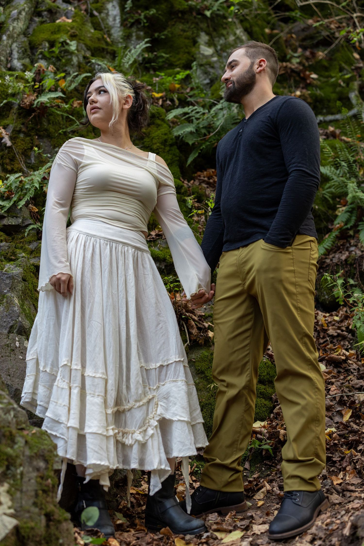 A couple in a forest setting, woman in white boho dress and man in black top with olive pants, holding hands.