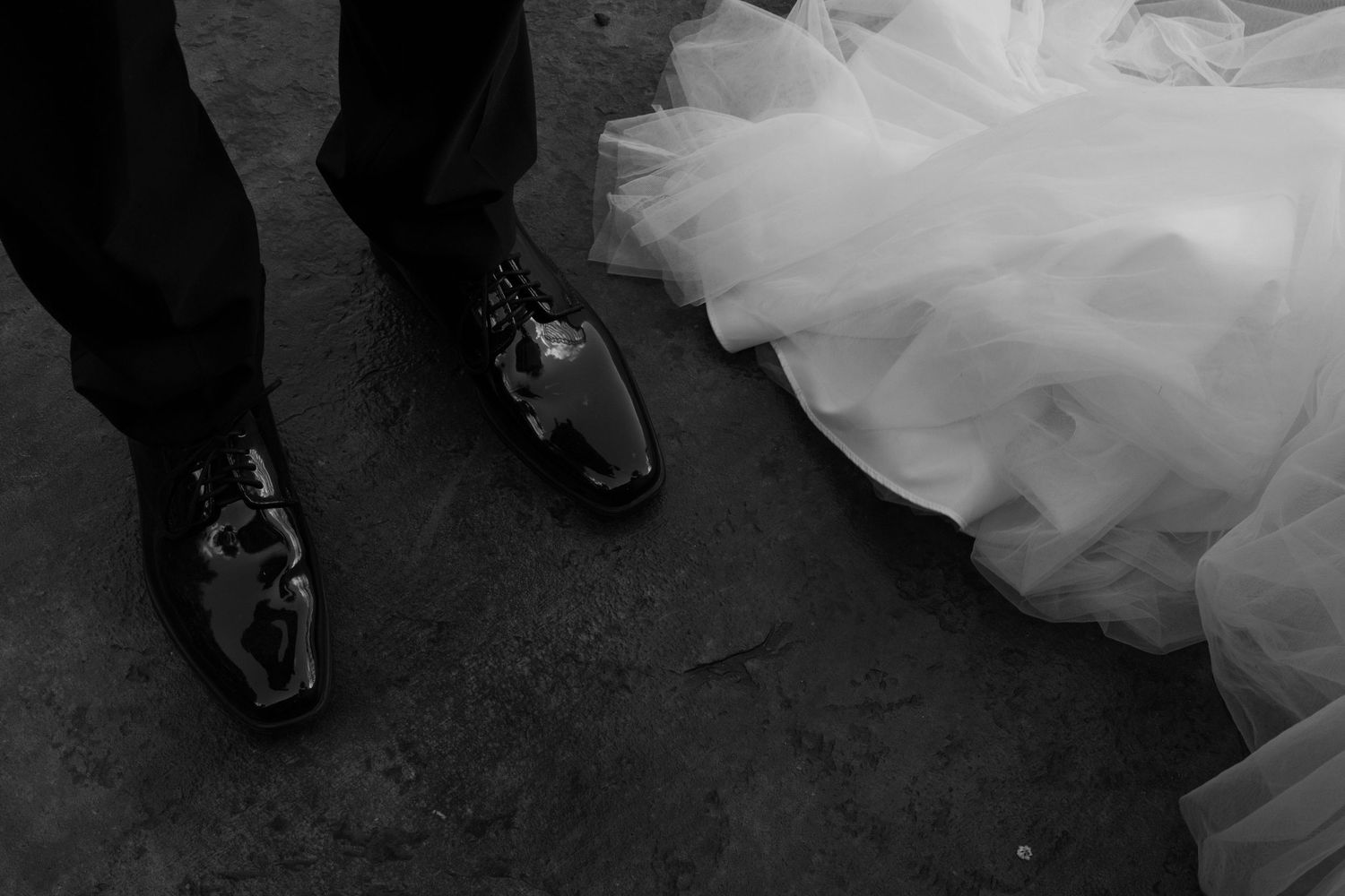 Black and white photograph showing the bottom of a wedding dress and dress shoes on a dark floor.