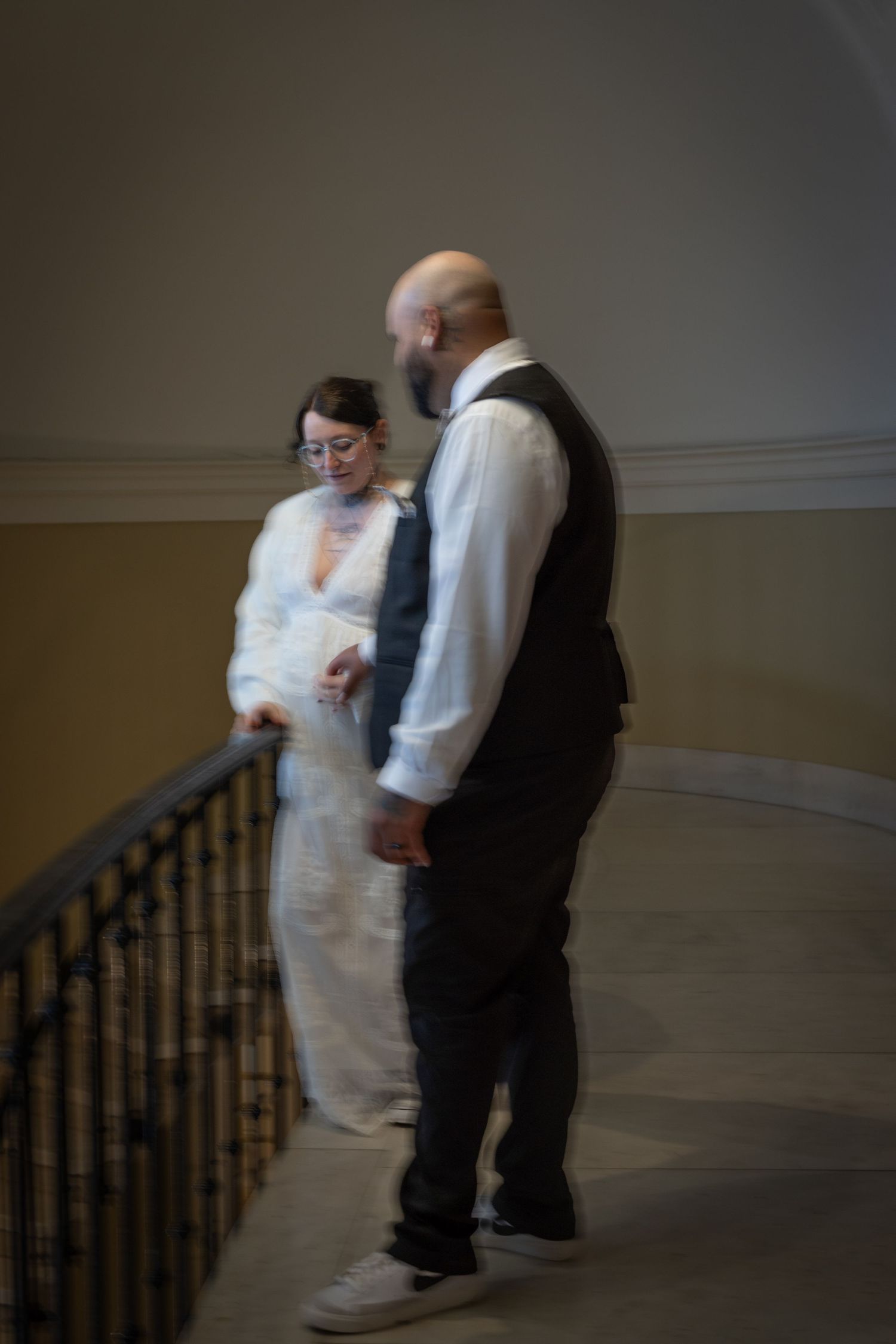A couple in wedding attire share an intimate Moment in a dimly lit hallway with wooden banisters. Milford Courthouse.